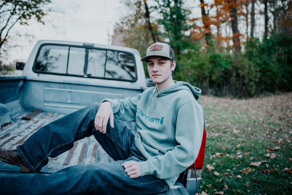 High school senior in casual wear sitting in the bed of a pickup truck surrounded by autumn trees.