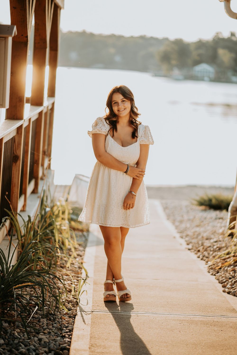 A high school senior in white dress poses on waterfront walkway at sunset.