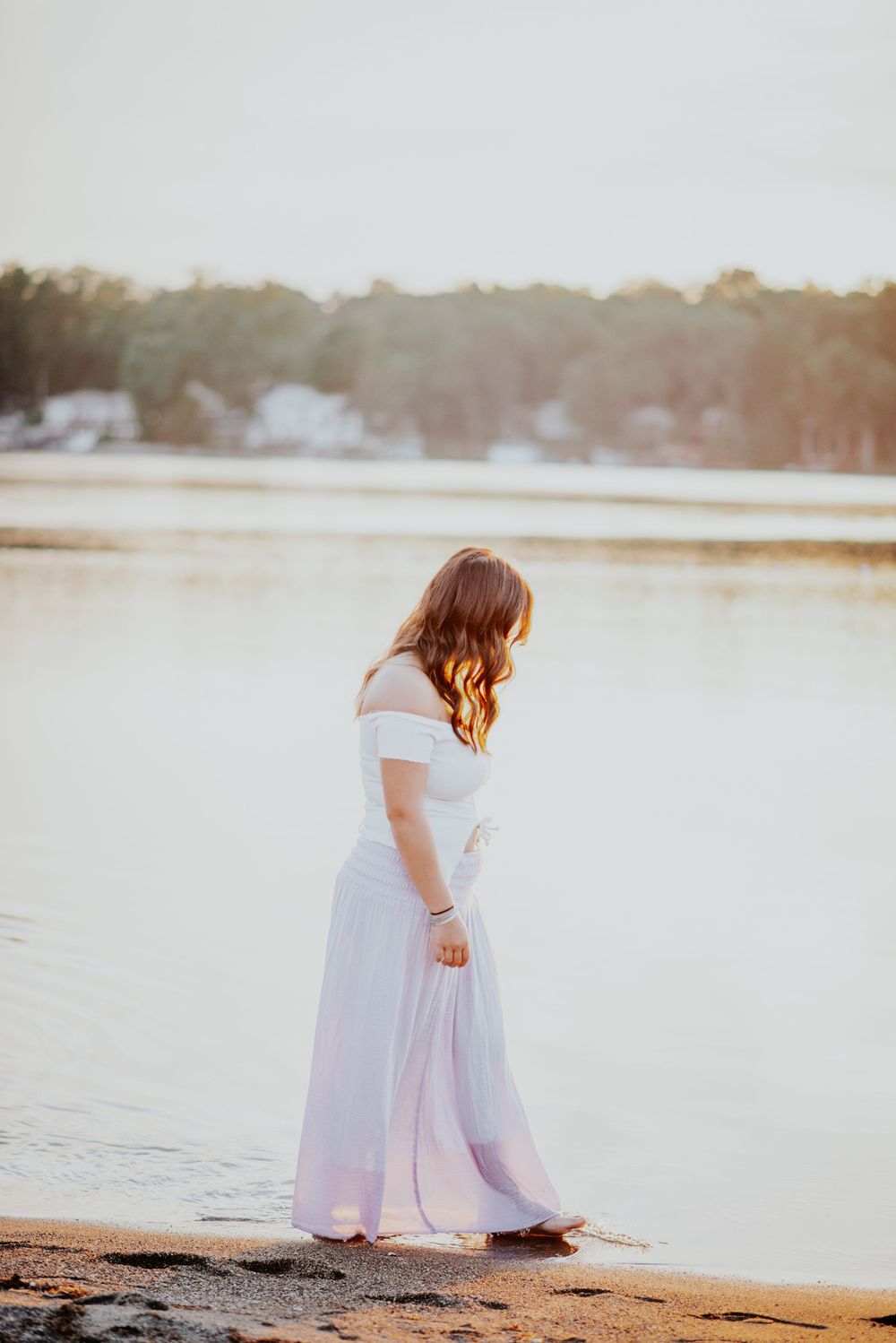 A high school senior in a flowing white dress stands contemplatively by a serene lake at sunset.