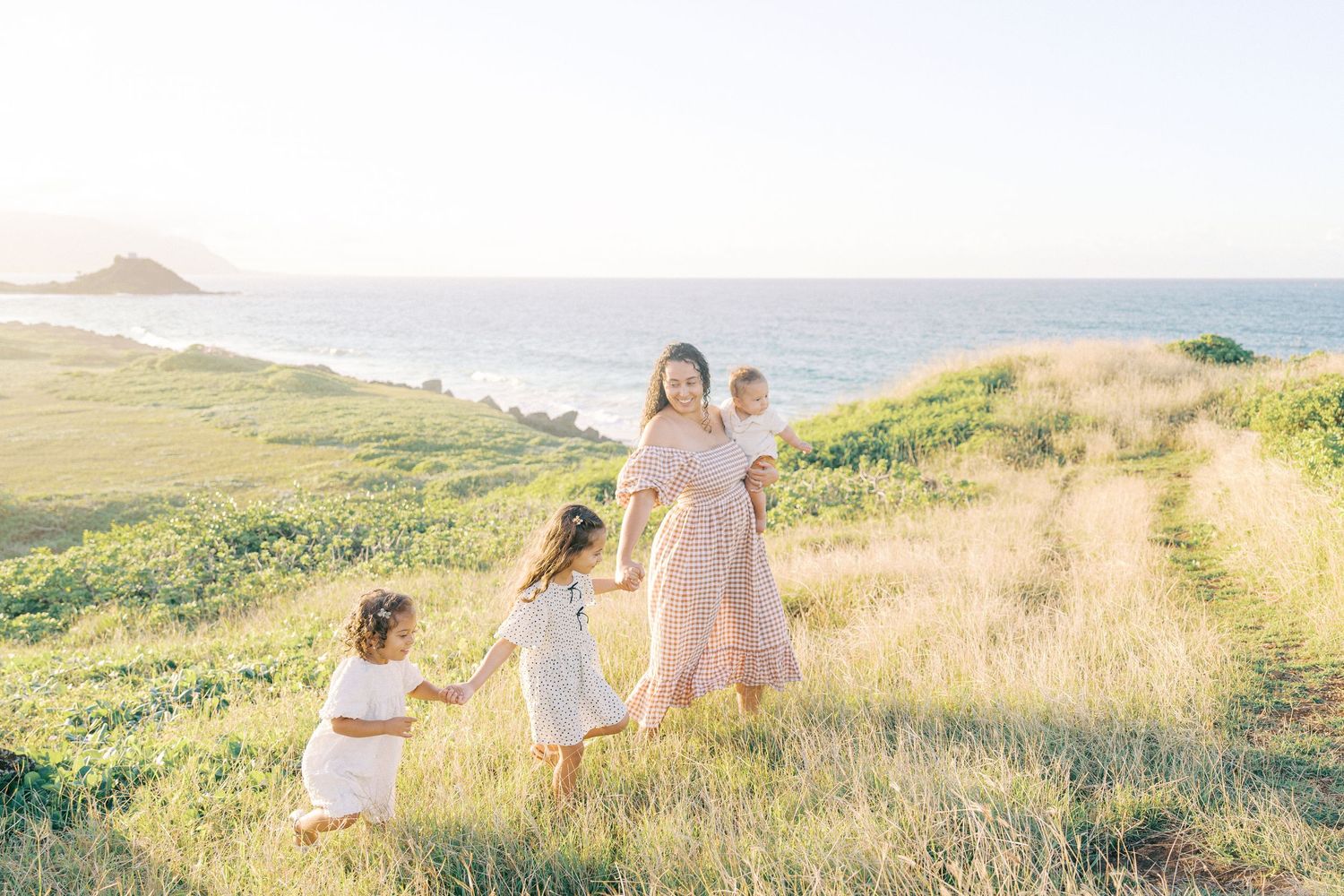 A family in light colored dresses walks together on grassy coastal path with ocean views.