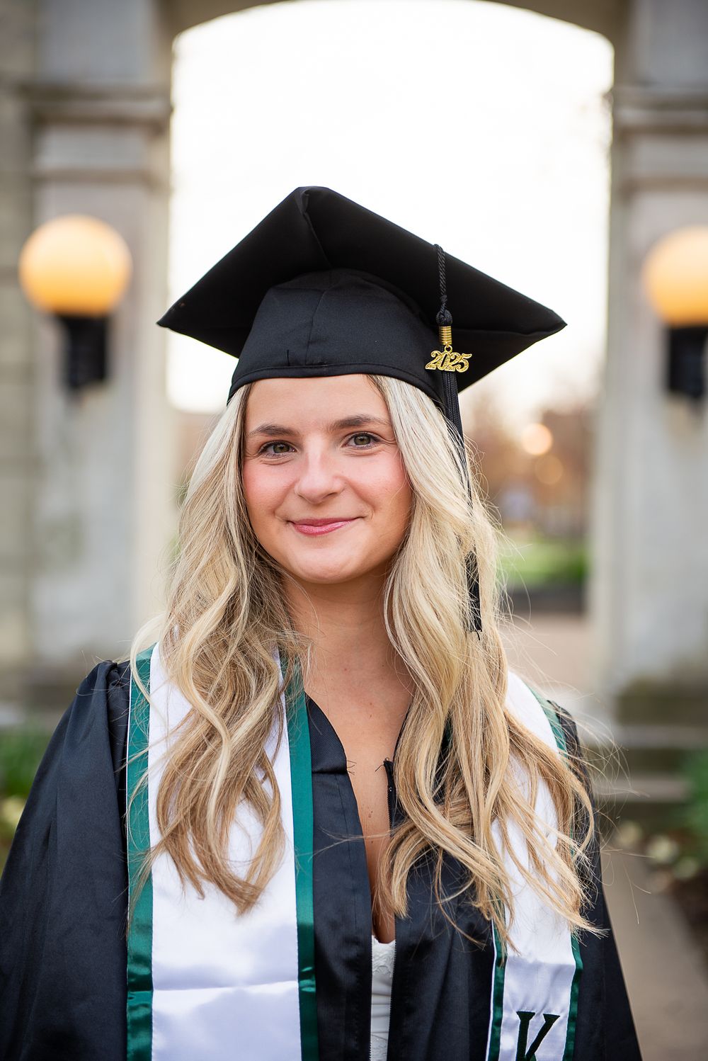 Graduate wearing cap and gown with stole poses under stone archway at sunset.