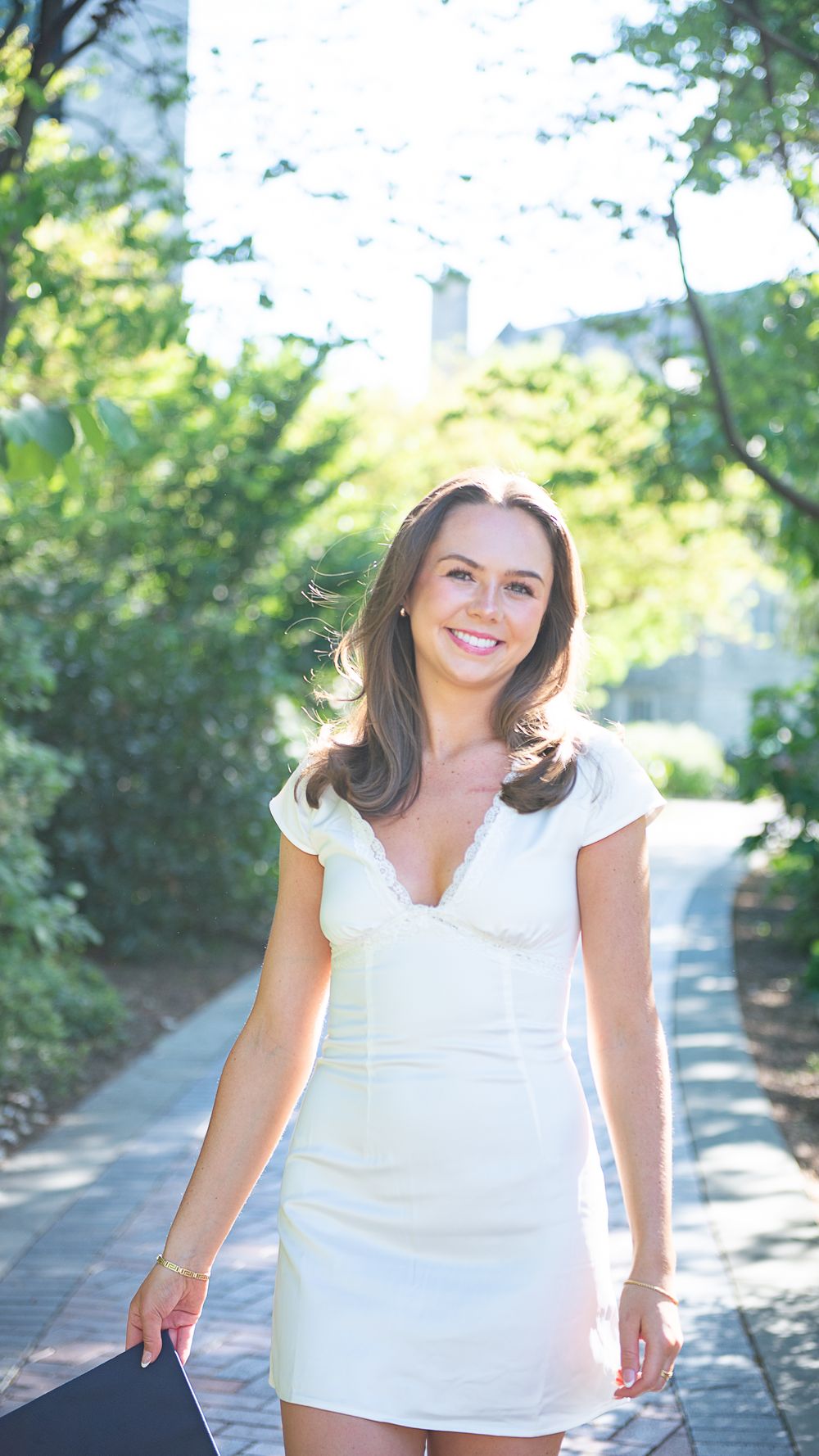 A graduate in a white summer dress poses on a sunlit garden path with trees in the background.