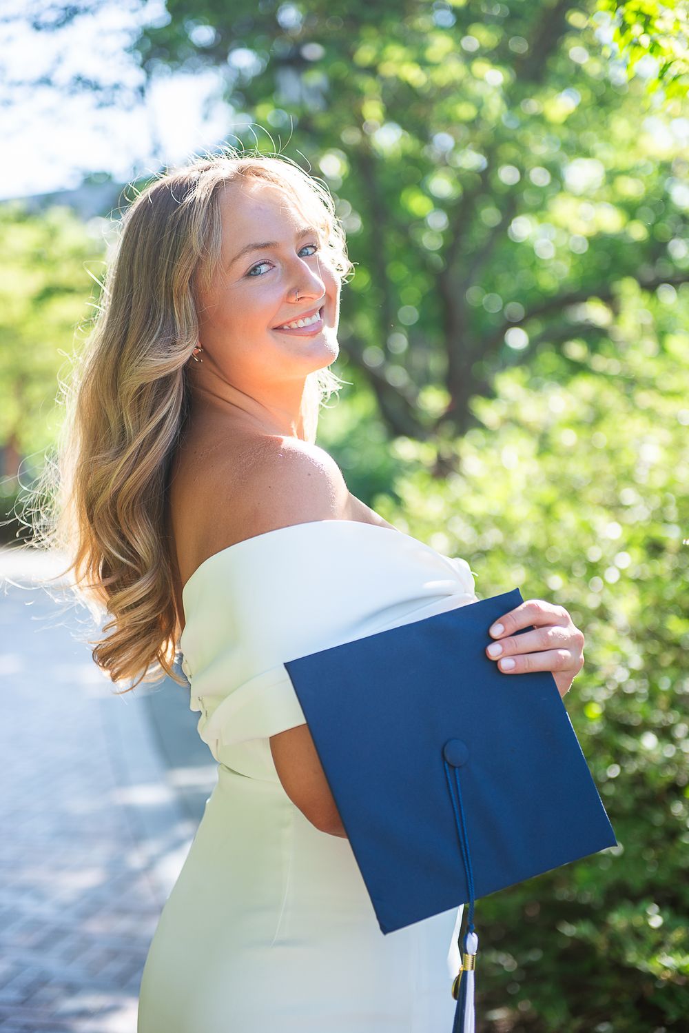 A graduate in white dress holds a blue mortarboard while posing outdoors in natural lighting.