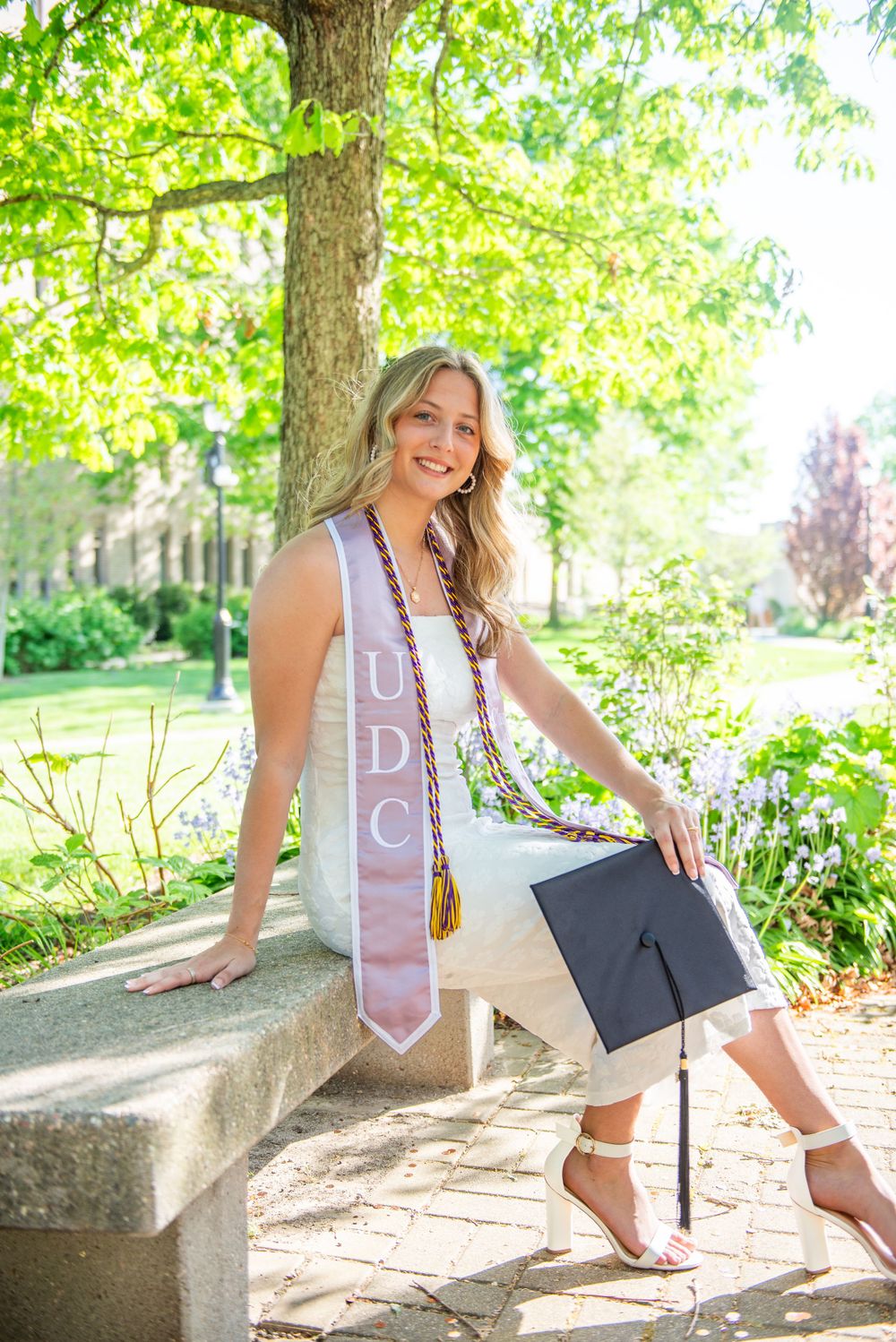 Graduate in white dress with sash poses under green trees during spring photo session.