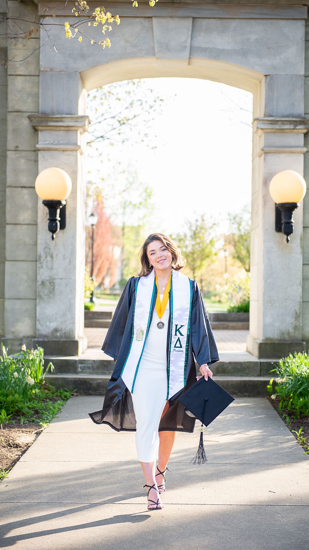 Graduate walking through stone archway in academic regalia holding graduation cap.