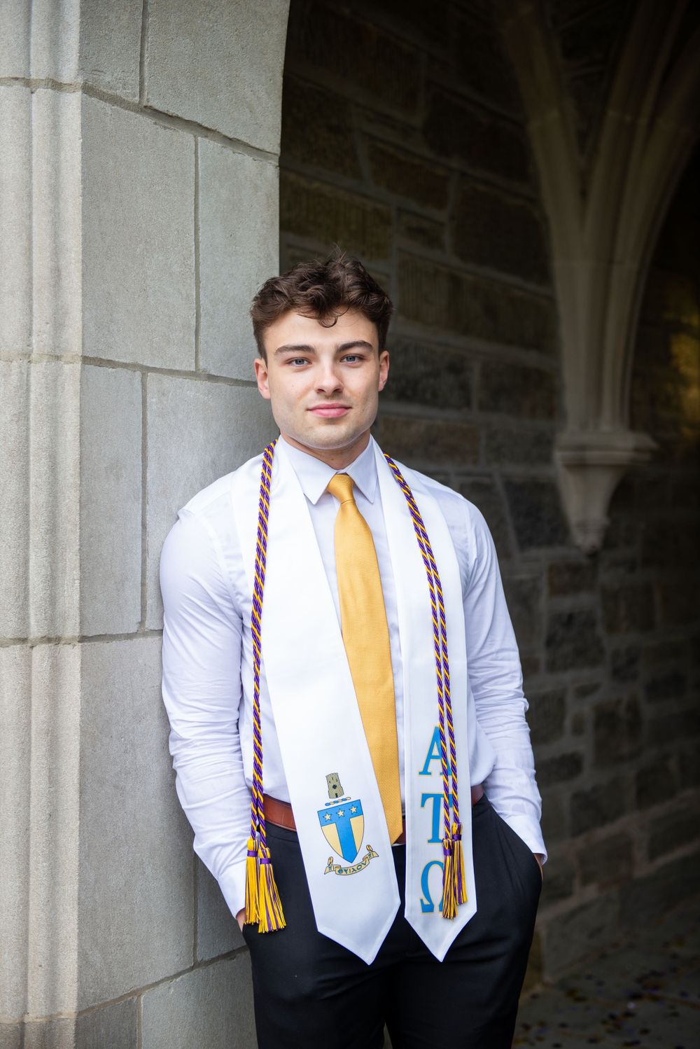 Graduate in formal attire with honor cords poses in collegiate Gothic archway.
