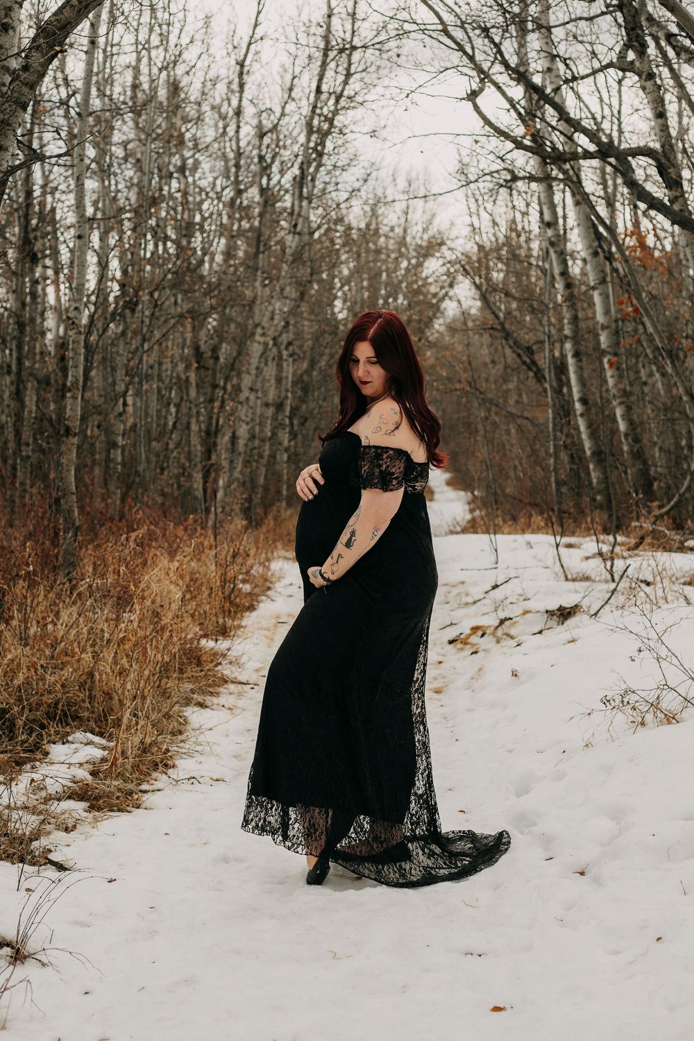 Pregnant woman in black lace off-shoulder gown poses gracefully on snowy path surrounded by bare winter trees.