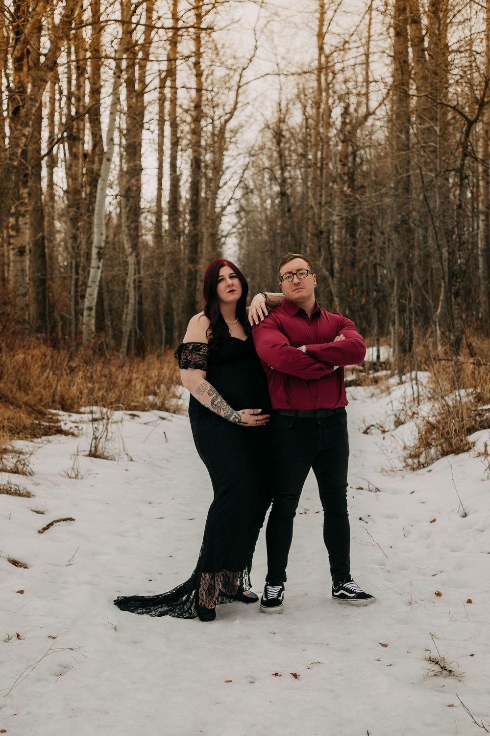 Couple posing in snowy winter forest, woman in black lace gown with tattoos, man in burgundy sweater and black jeans.