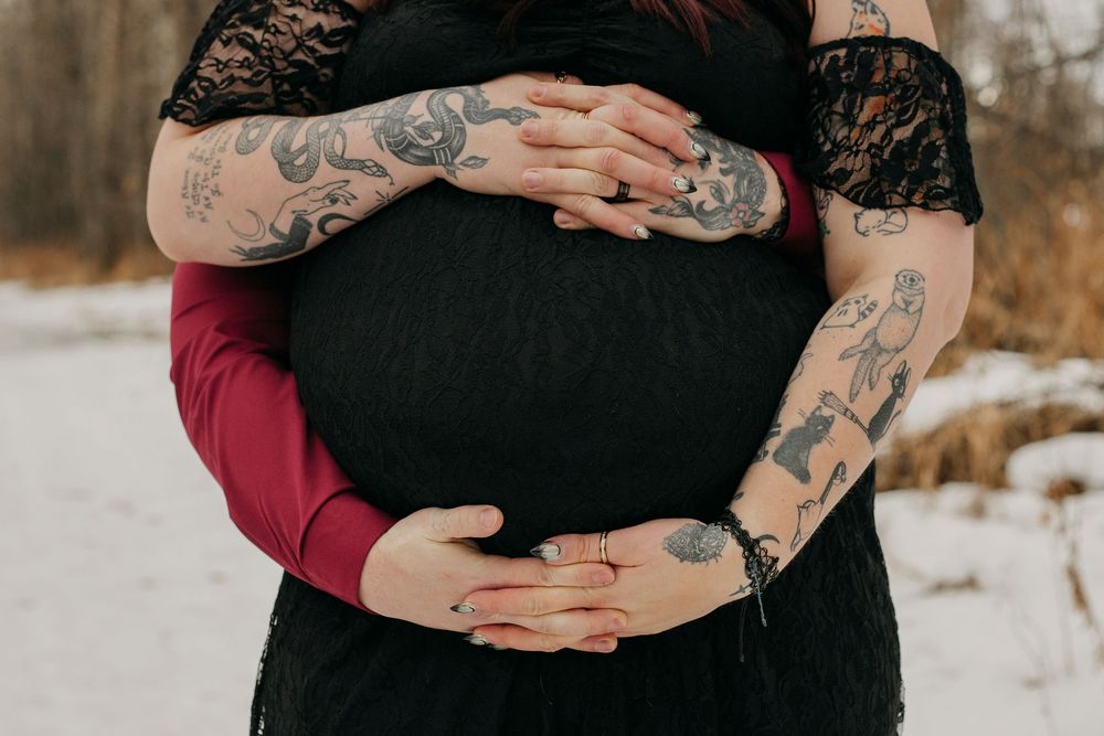 Pregnant woman in black lace dress with tattooed arms cradling baby bump in snowy outdoor maternity photo.