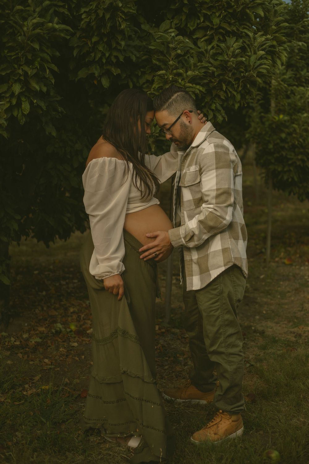 A romantic maternity photo shows a couple embracing in an orchard with soft vintage-style lighting