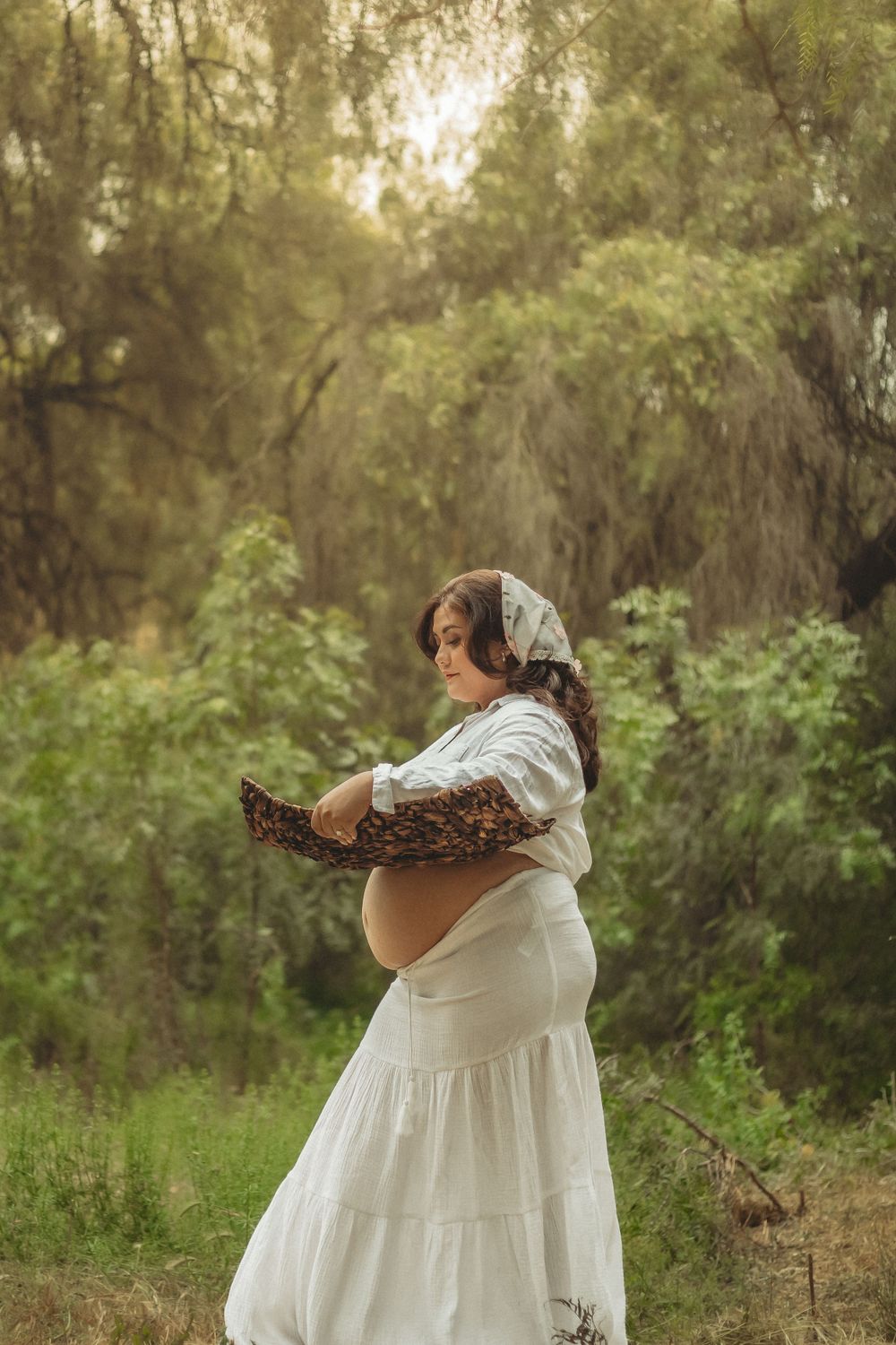 Ethereal maternity photo in a white flowy dress holding a woven basket in golden sunlit forest setting