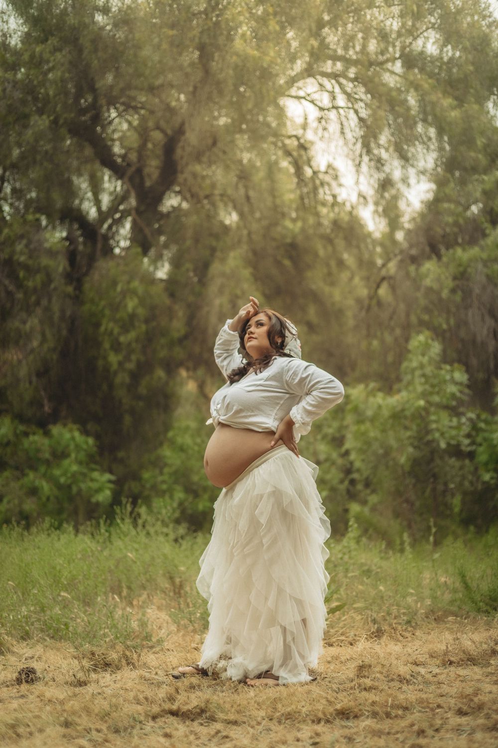 An expectant mother in a flowing white skirt and top poses gracefully in a misty woodland setting during golden hour in Glendora California