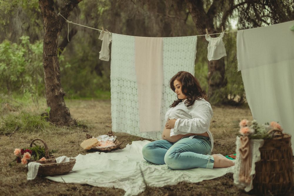 Mommy relaxes on a blanket outdoors with white linens hanging on clothesline for a romantic vintage maternity photography session