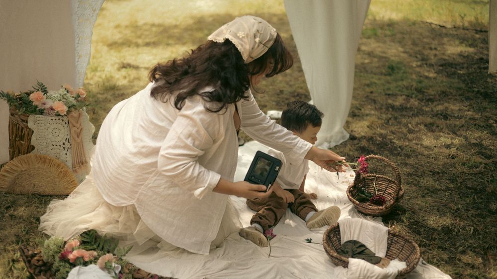 Pregnant mother in a white dress sits on the ground sharing polaroids with her child