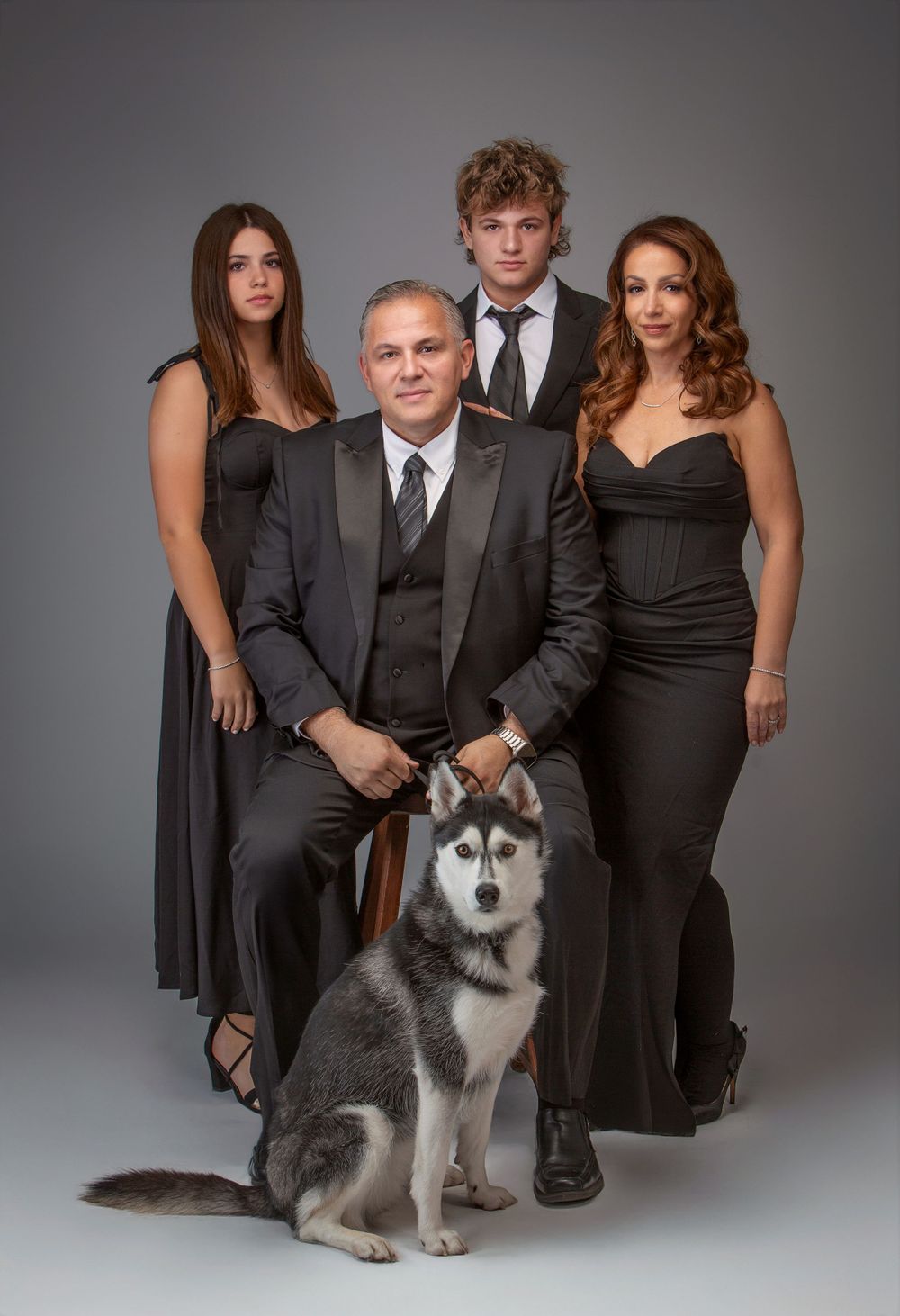 Studio portrait of four people in elegant black formal attire with a beautiful husky dog in front on grey background.