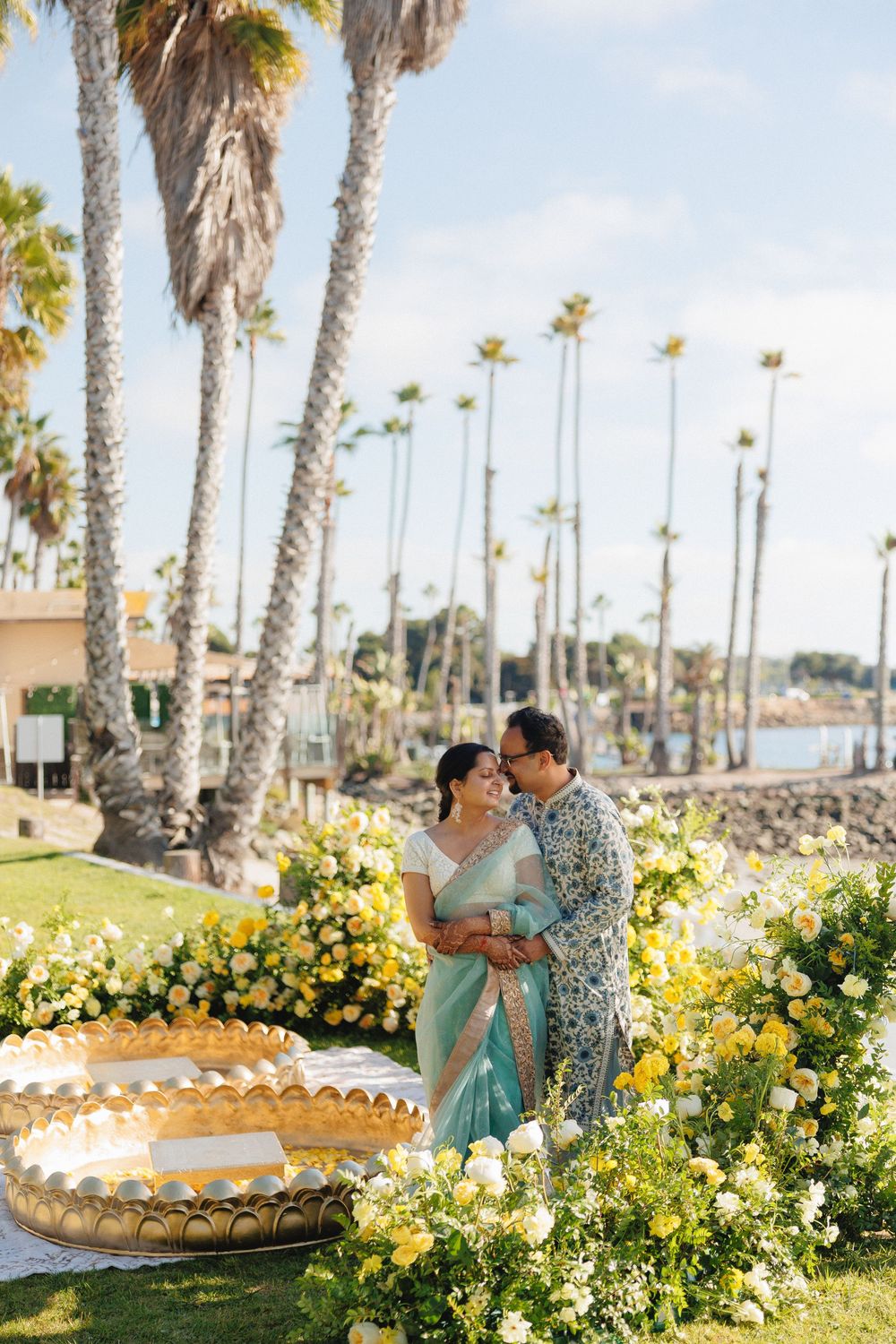 A couple embraces among yellow flowers and palm trees at a beachside wedding venue in California.