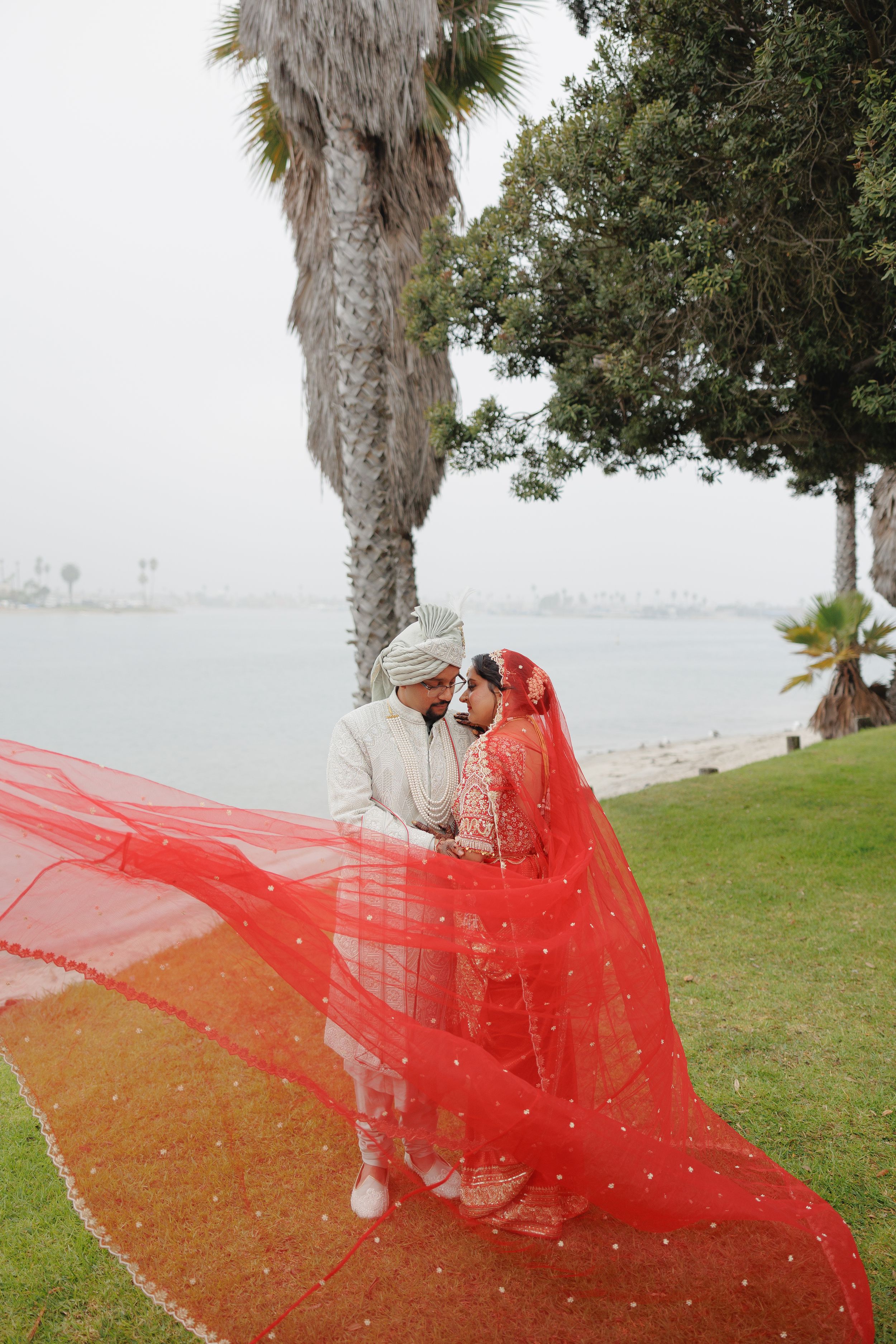 A flowing red veil billows in the wind along a palm tree-lined coastal path.