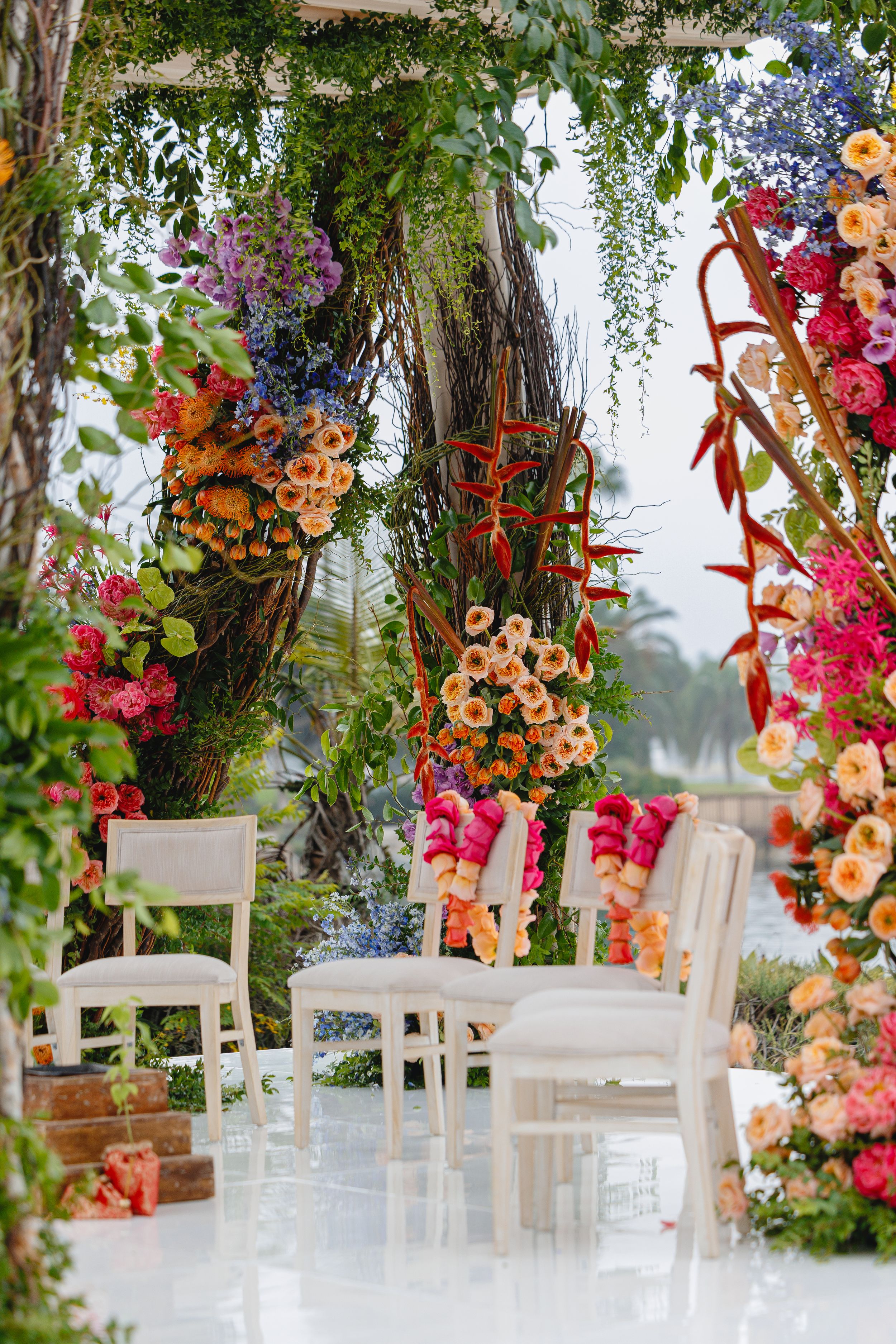 Elegant outdoor wedding ceremony setup with white chairs and vibrant pink and orange floral arrangements hanging from trees.