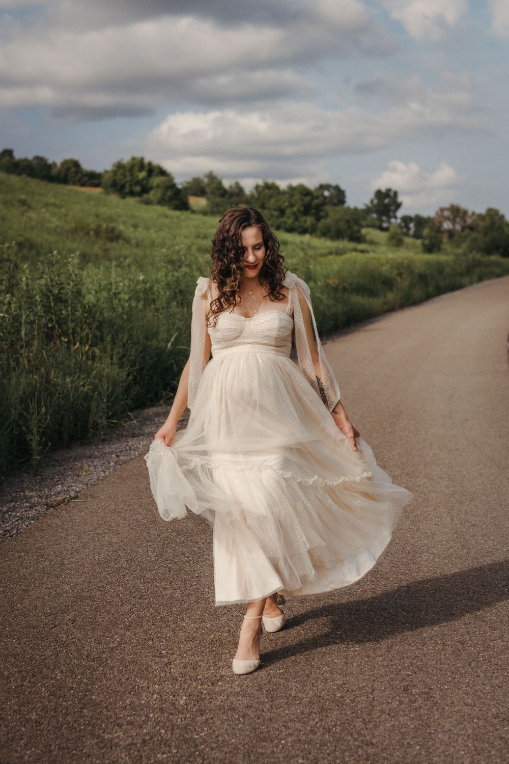 An expecting mother in a flowing white tulle gown walks down a country road, her dress billowing in the breeze at Seven Islands State Park.
