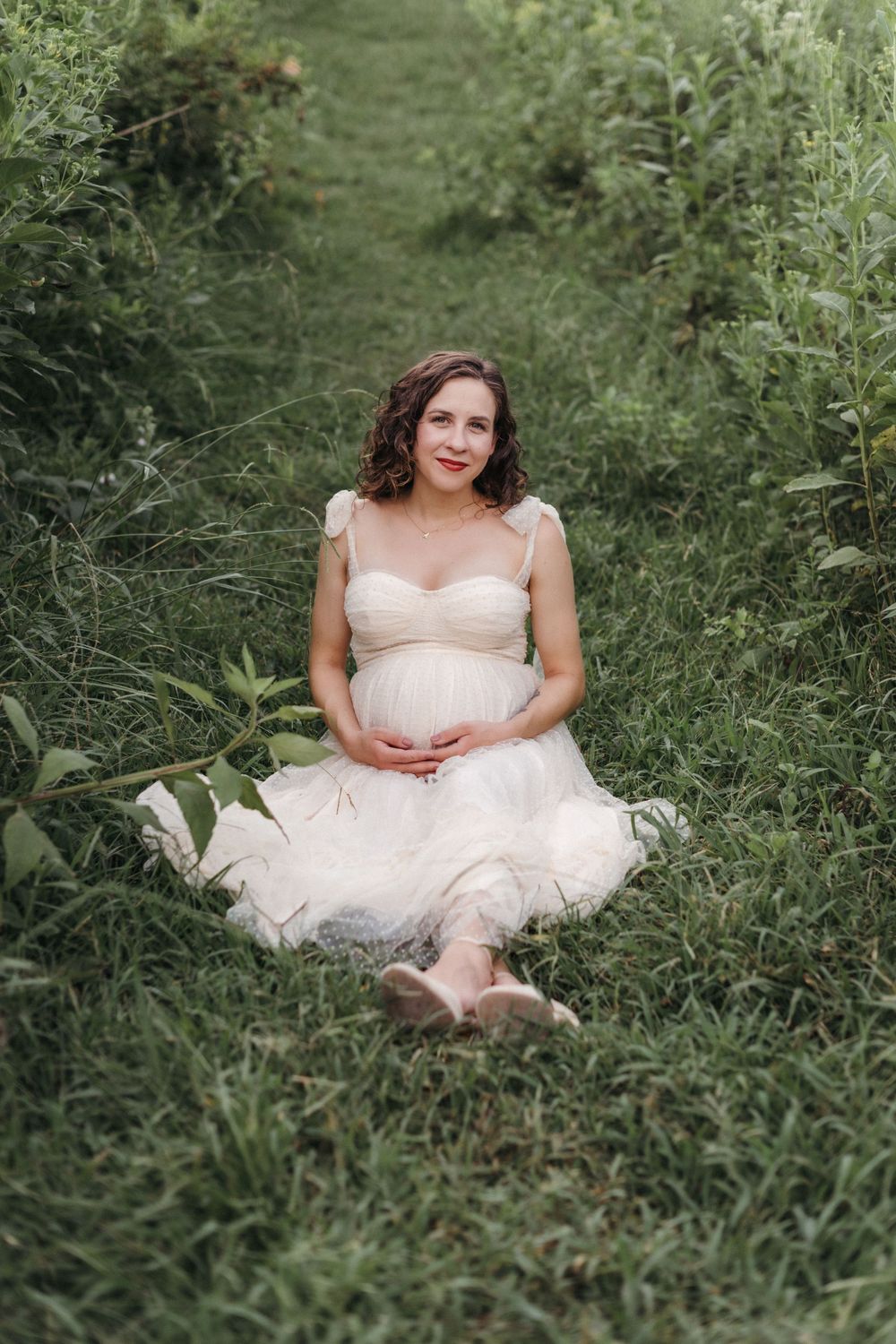 Pregnant woman in white dress sitting on grass path in lush green garden, cradling baby bump during maternity photo shoot at Seven Islands.