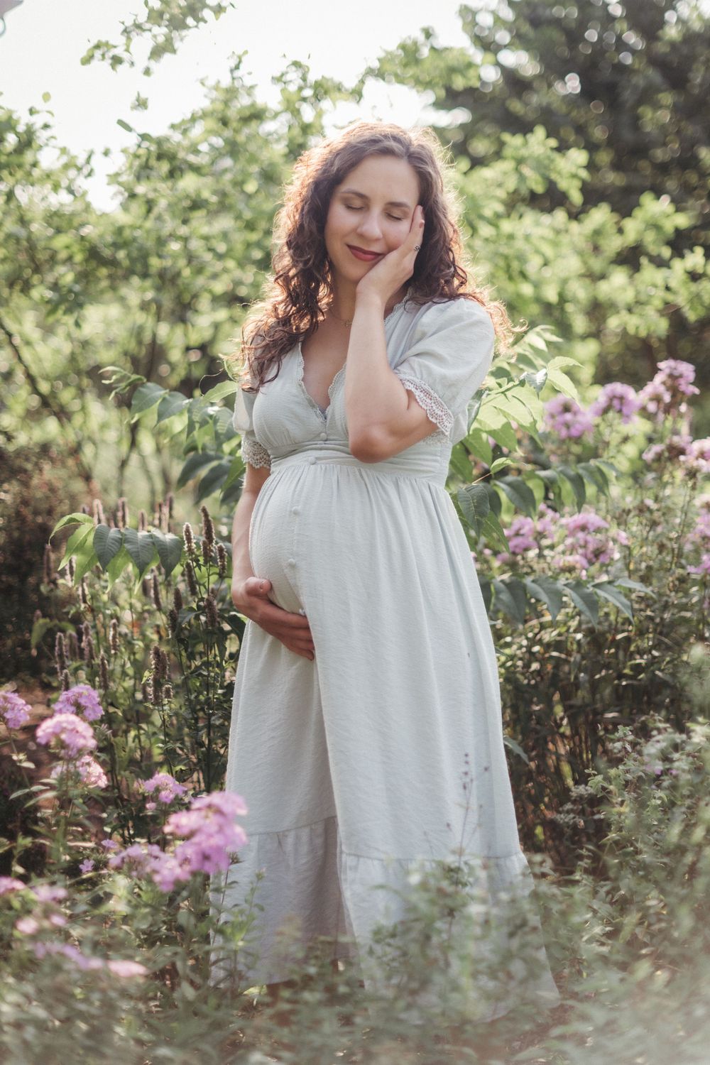 Pregnant woman in white dress cradling her baby bump eyes closed surrounded by purple flowers in a lush garden at Seven Islands State Park.