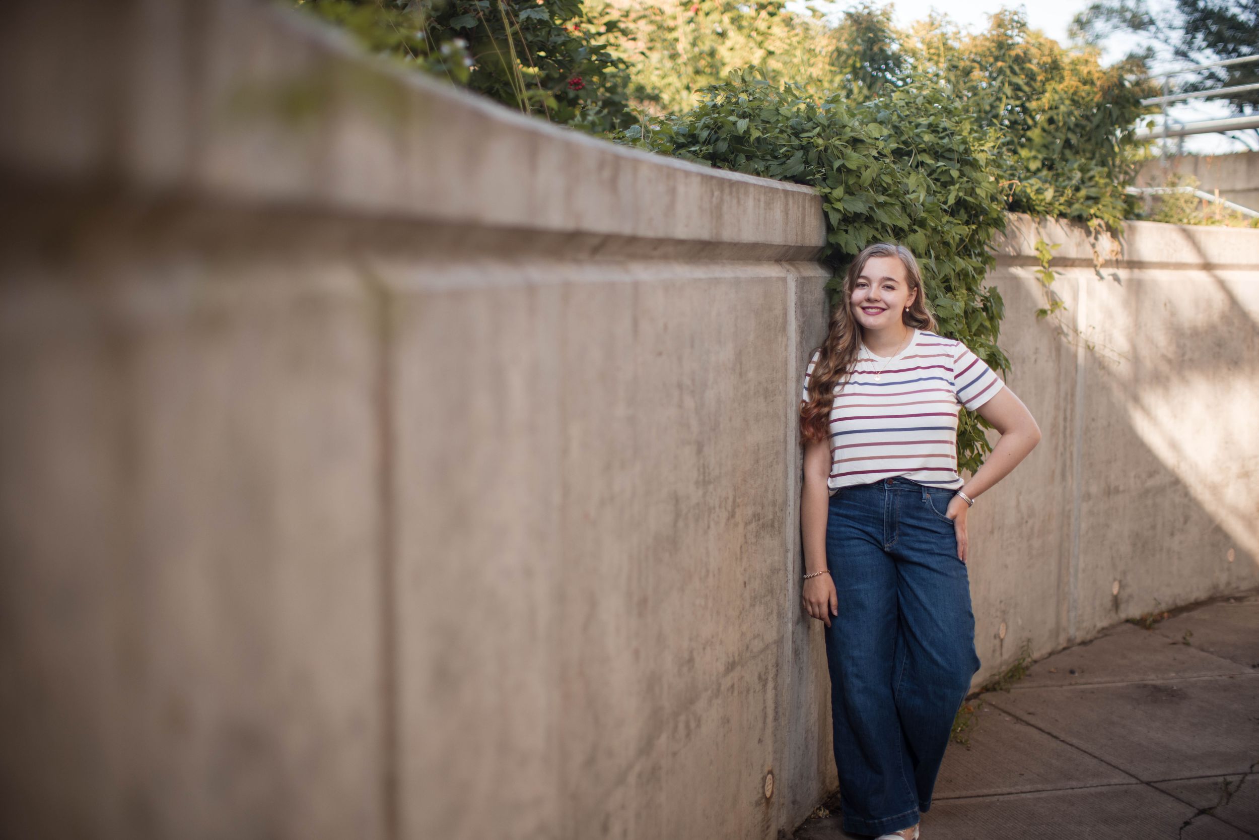 Person in striped shirt and jeans leans against concrete wall in outdoor urban setting with natural lighting.