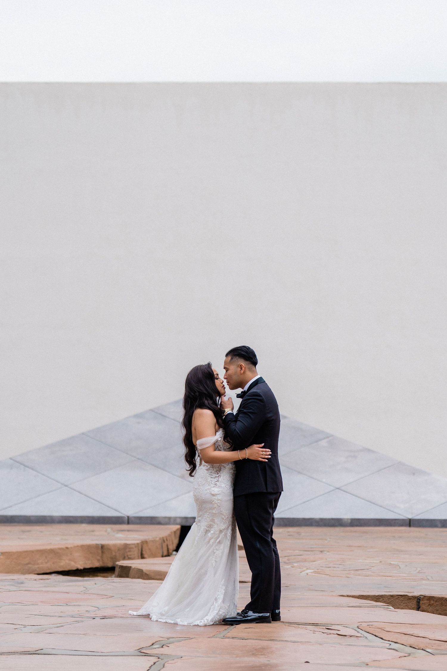 A bride and groom share a romantic kiss in front of a white wall, the bride in a fitted gown and groom in a black tuxedo.