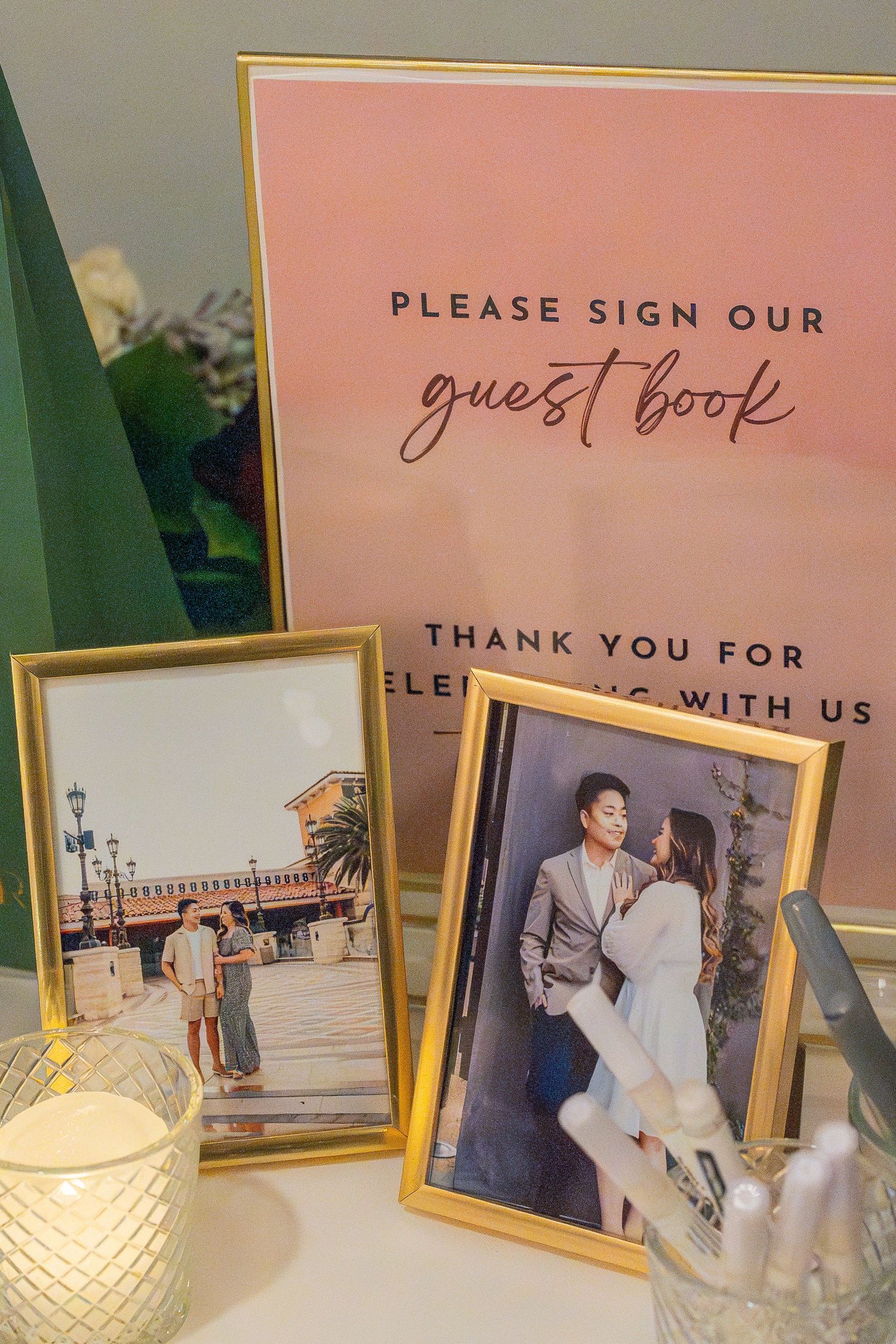 Wedding guest book sign display with gold-framed couple photos, candle, and pens on elegant white table.