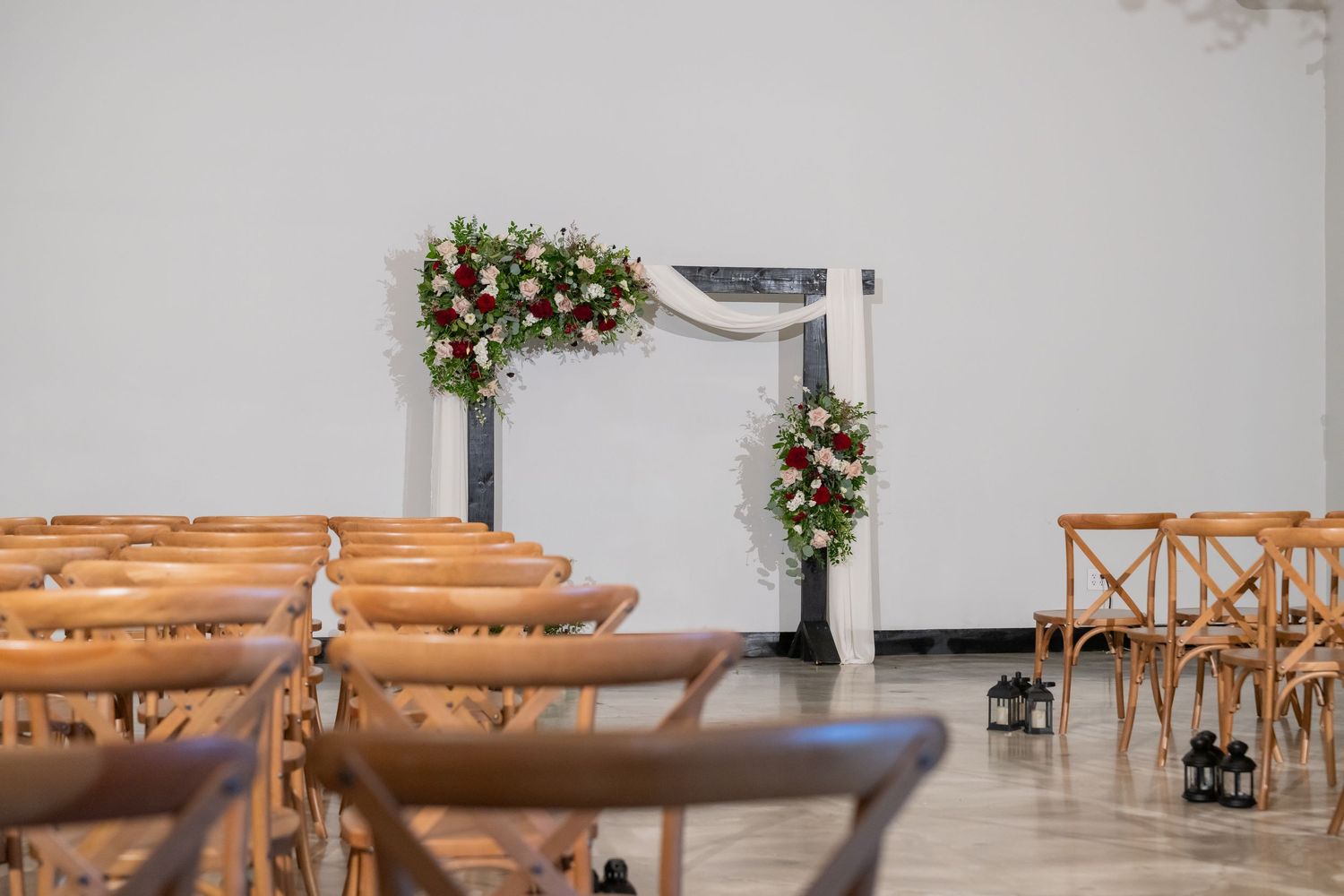 Elegant wedding ceremony setup with wooden chairs, floral arch adorned with red and white flowers against a white wall.