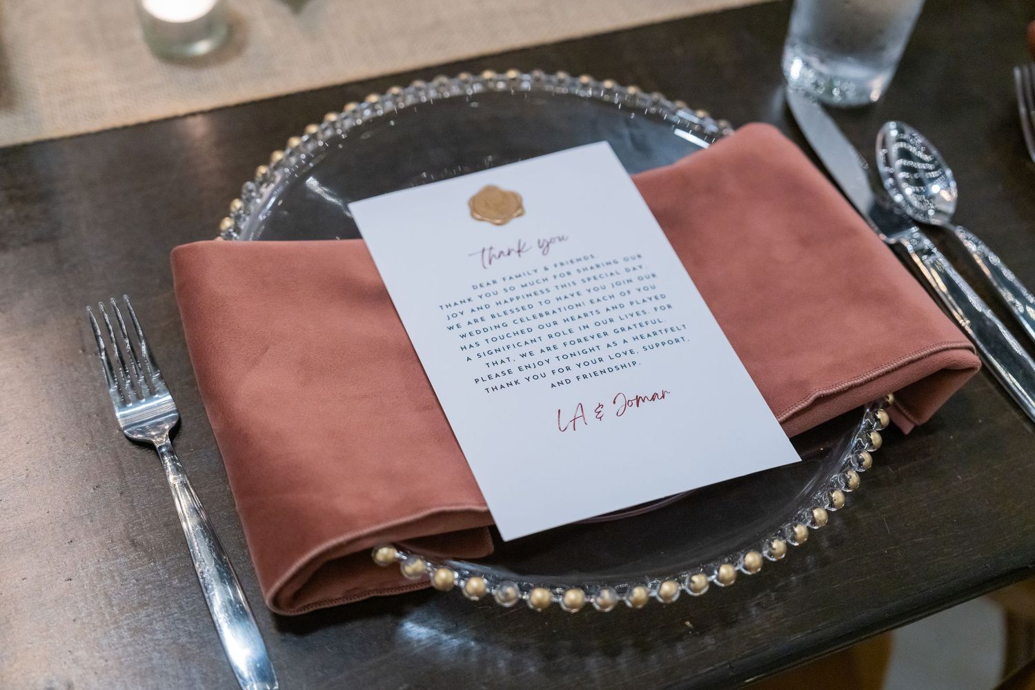 Elegant wedding place setting with dusty rose napkin, beaded charger plate, and personalized thank you card.