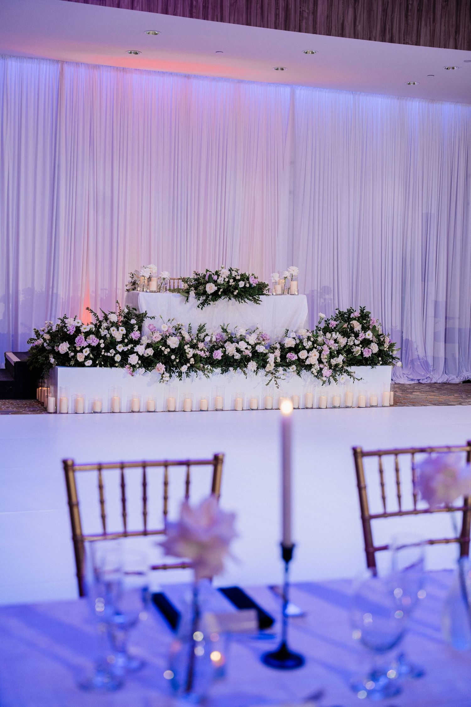Elegant wedding reception venue with floral arrangements, white draping, candles, and Chiavari chairs in soft purple lighting.