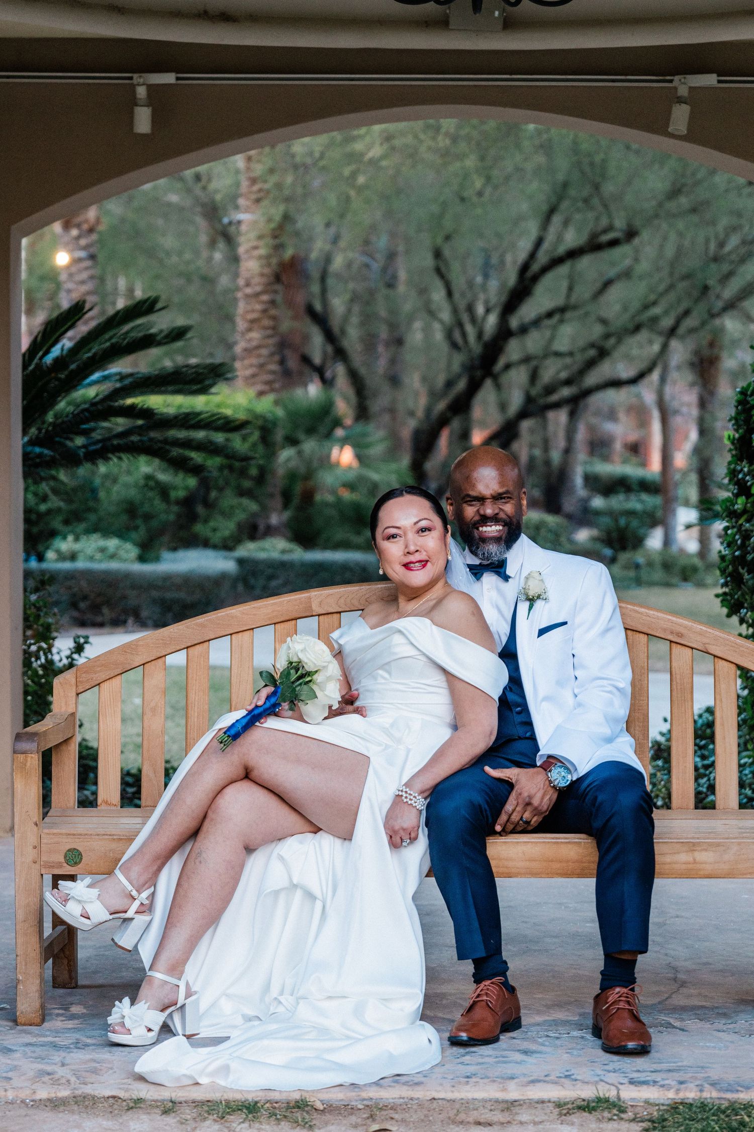 A bride and groom in wedding attire sit together on a wooden bench in a lush garden setting.