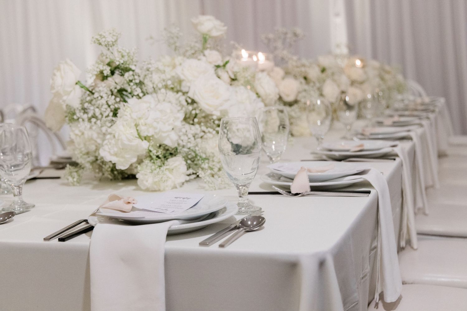 Elegant white wedding reception table with floral centerpieces, silver cutlery, and white linens in a bright venue.