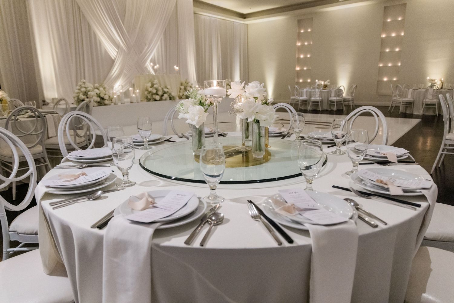 Elegant white wedding reception table setting with glass lazy Susan centerpiece, white linens, and crystal glasses in a draped ballroom.