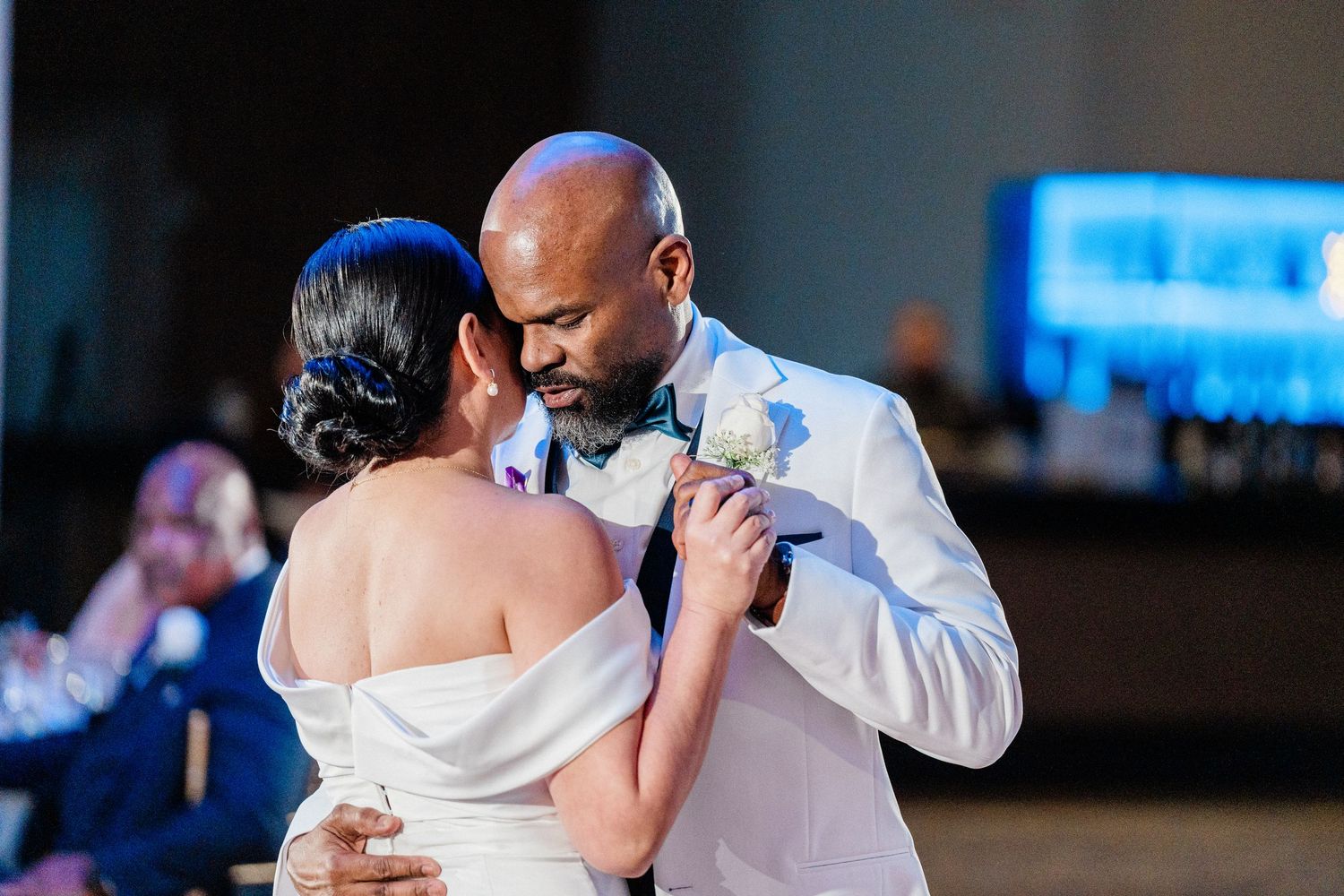 Bride and groom share their first dance at wedding reception, groom in white suit with boutonniere.