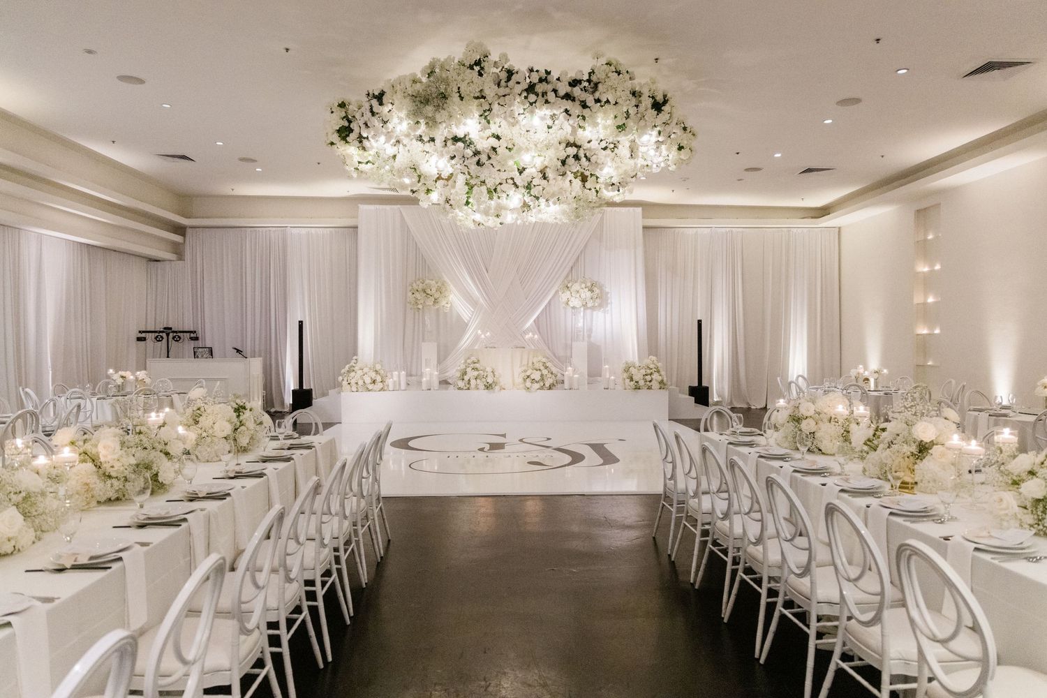 Elegant all-white wedding reception hall with floral chandelier, draped walls, long tables, and monogram dance floor.