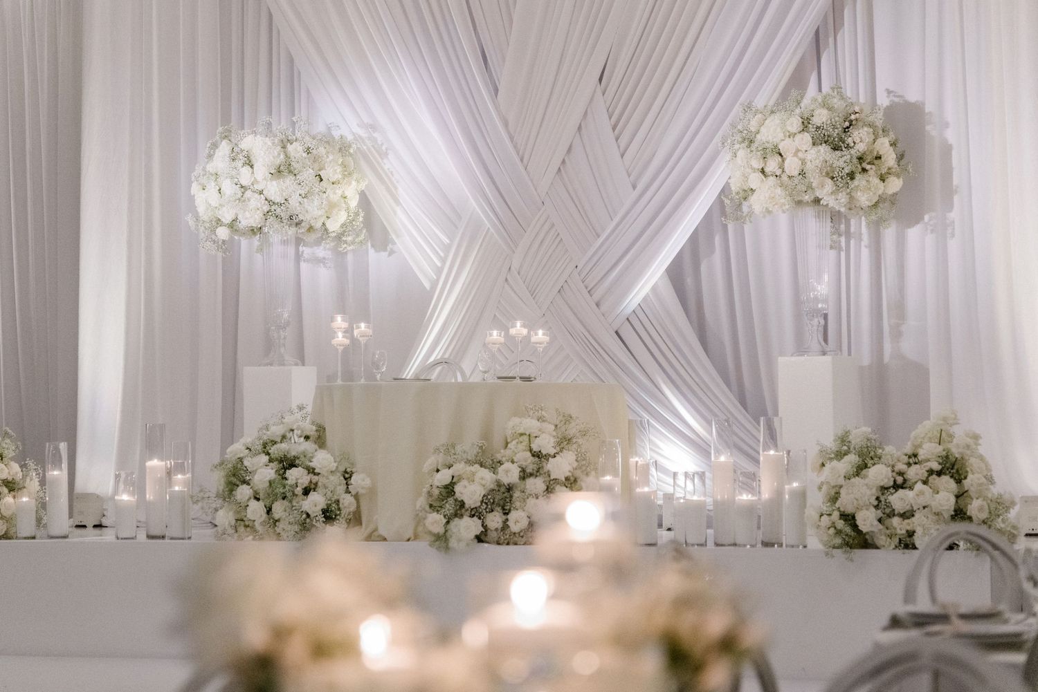 Elegant all-white wedding sweetheart table with floral arrangements, draped fabric backdrop, and glowing candles.