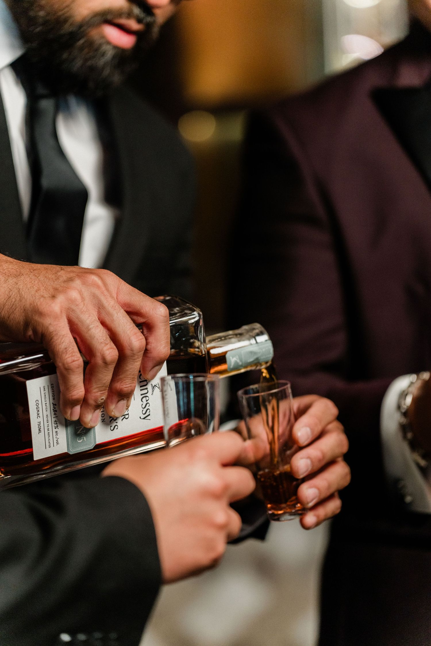 Man pouring Hennessy cognac into shot glasses at an upscale event with men in suits nearby.