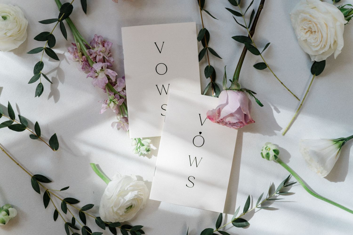 Two elegant wedding vow booklets surrounded by fresh white roses, pink flowers, and green eucalyptus on a white background.