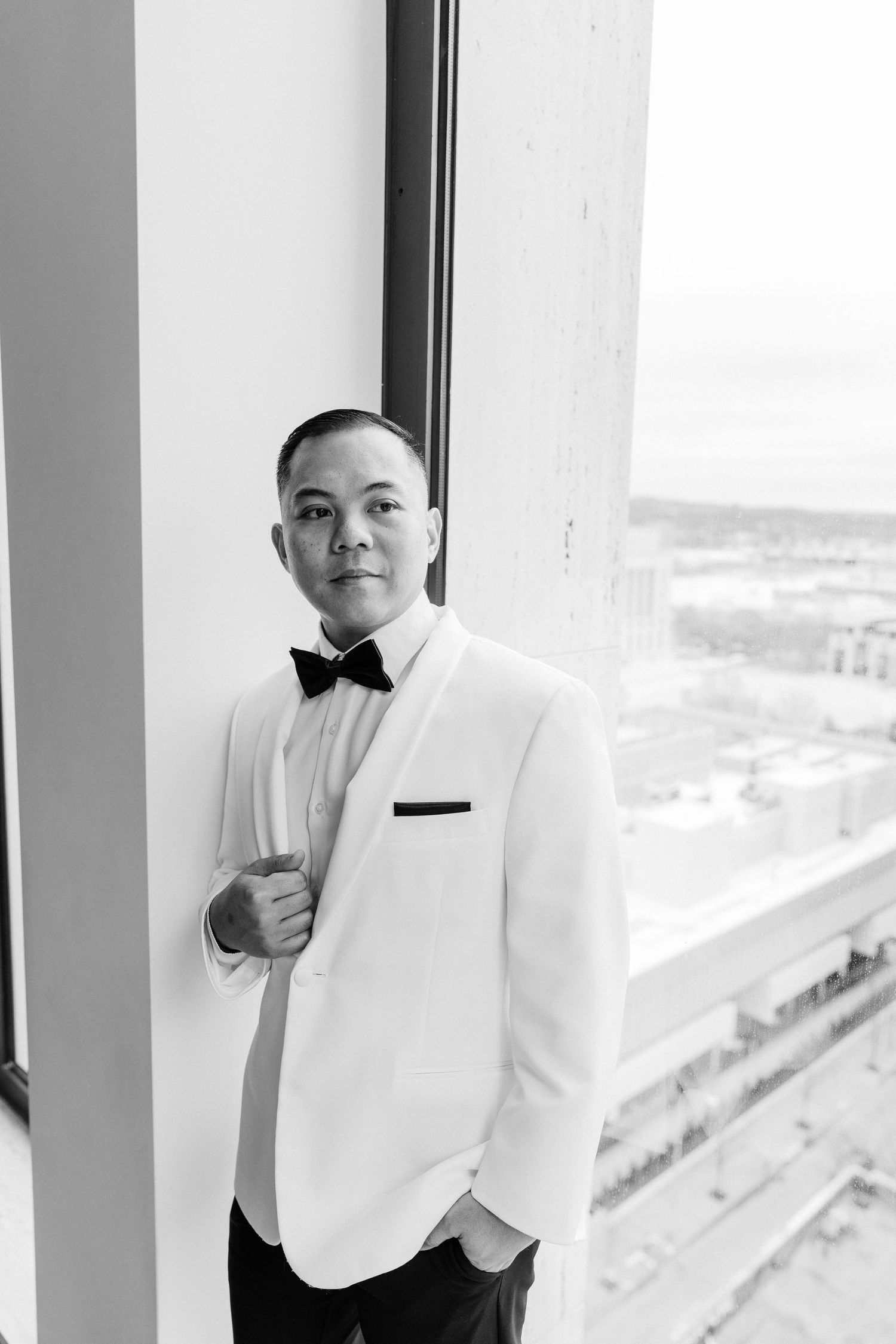 Black and white portrait of a groom in a white tuxedo jacket and bow tie standing by a large window.