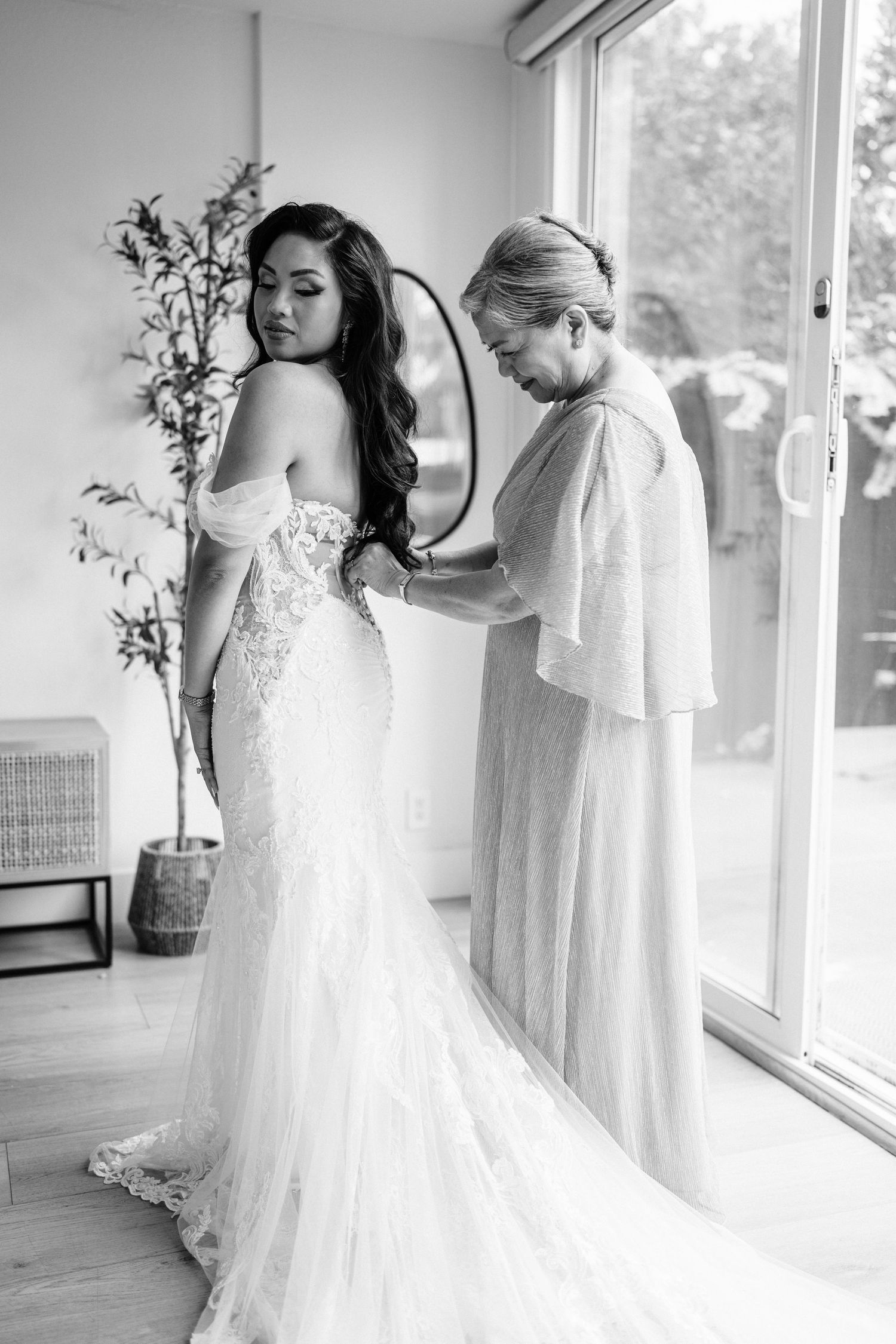 Black and white photo of a bride in a lace mermaid gown being helped with her dress by an older woman near a window.