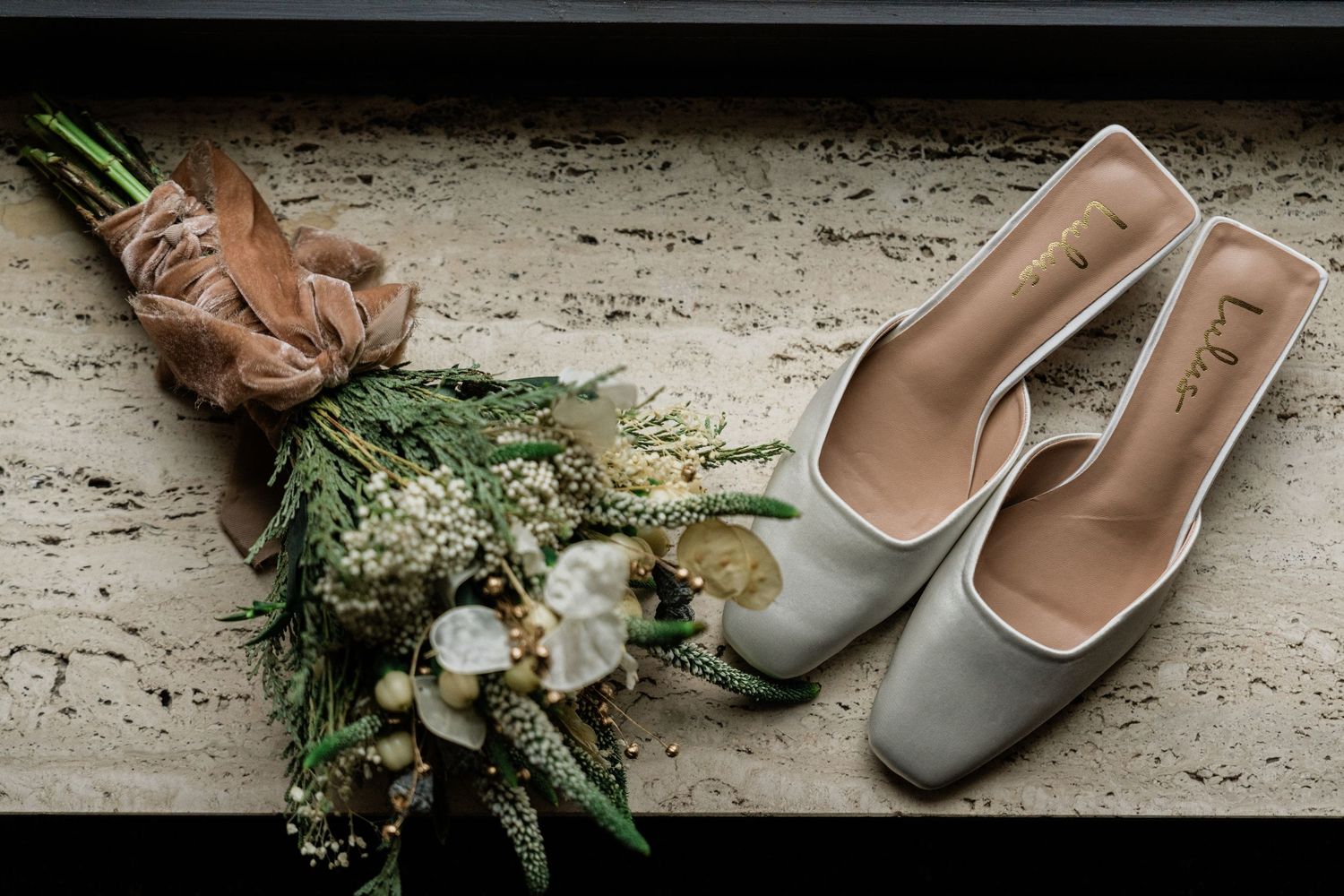 Elegant grey kitten heel mules beside a rustic bridal bouquet with greenery and white flowers on a stone surface.