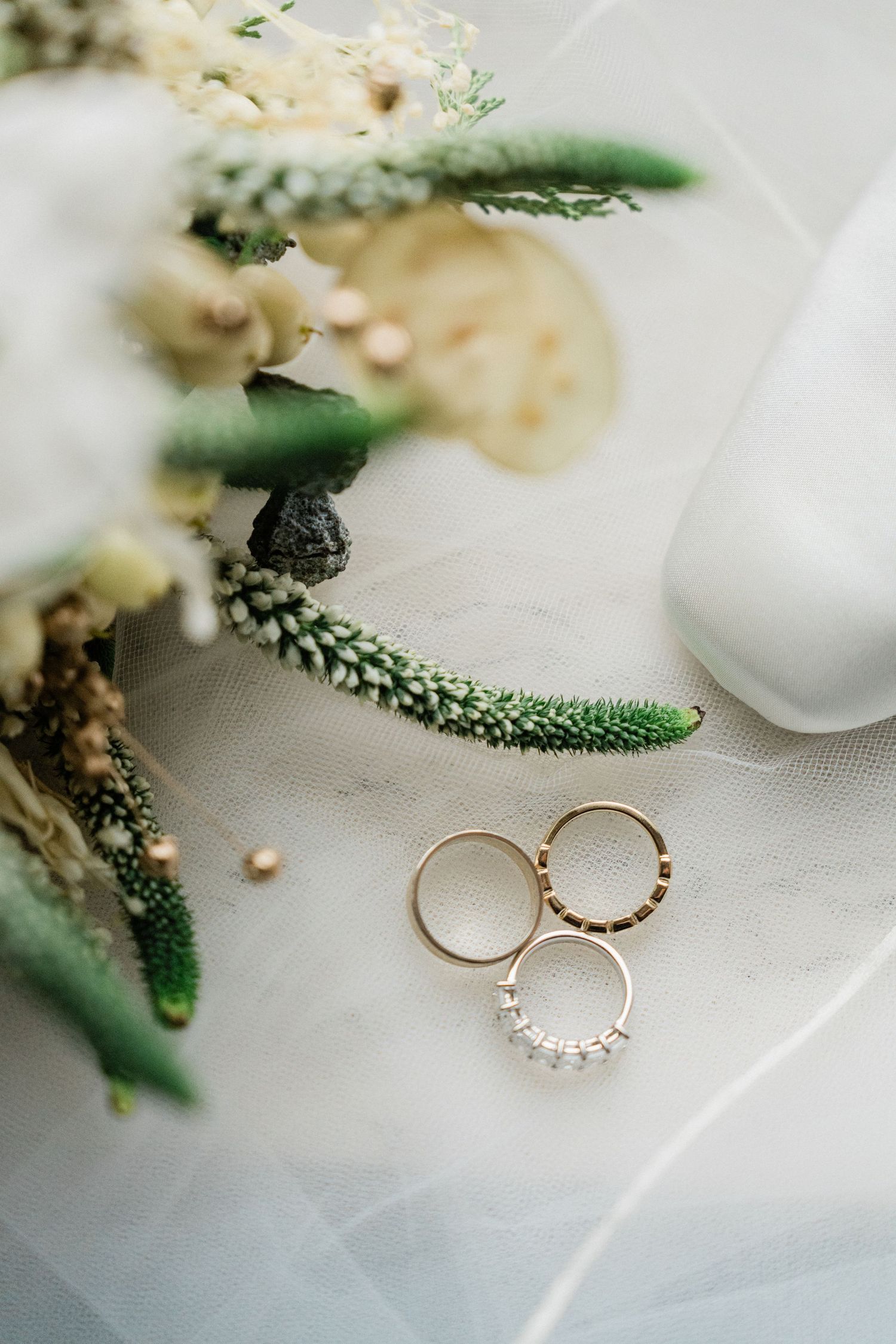 Gold wedding rings with diamonds beside a bridal bouquet of white and green flowers on a soft white veil.