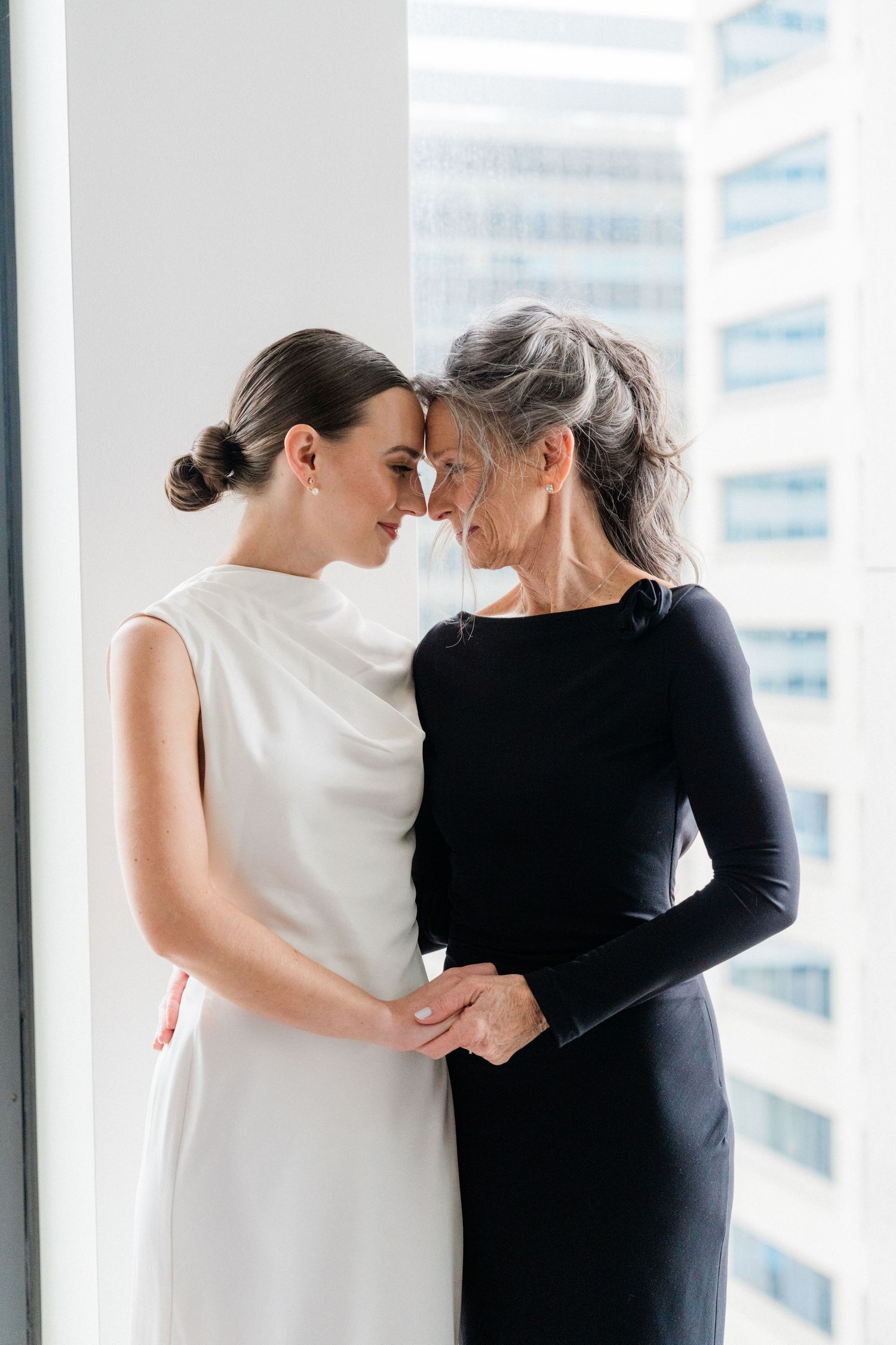 Two women in elegant black and white dresses share an intimate moment, touching foreheads tenderly near a bright window.