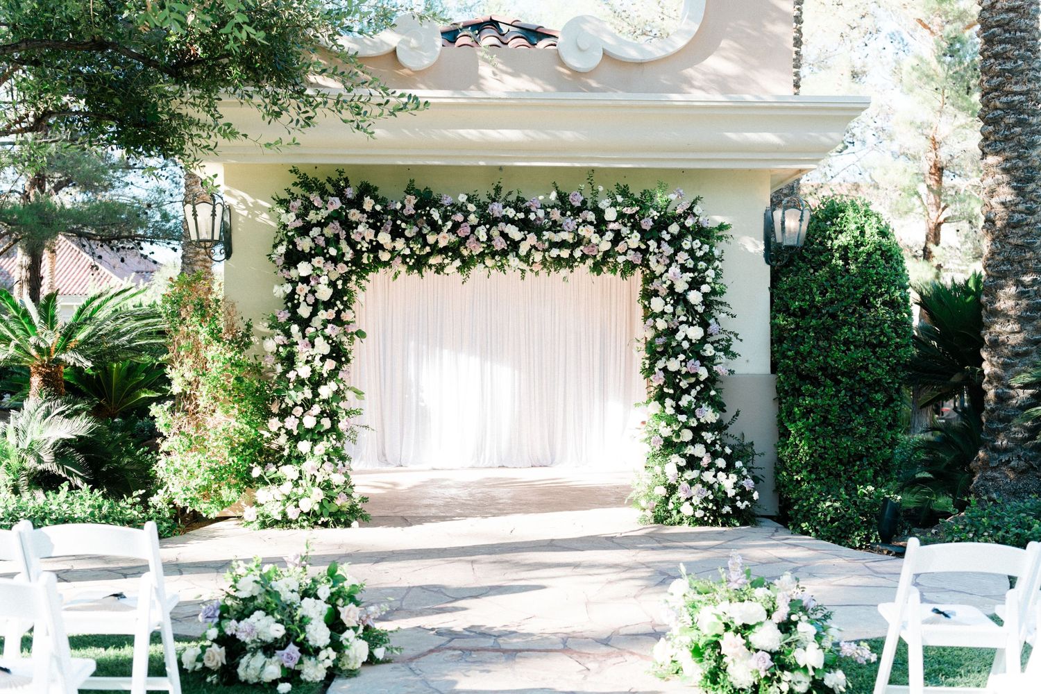 Elegant outdoor wedding ceremony arch adorned with lush white floral arrangements, sheer draping, and white garden chairs on a stone patio.