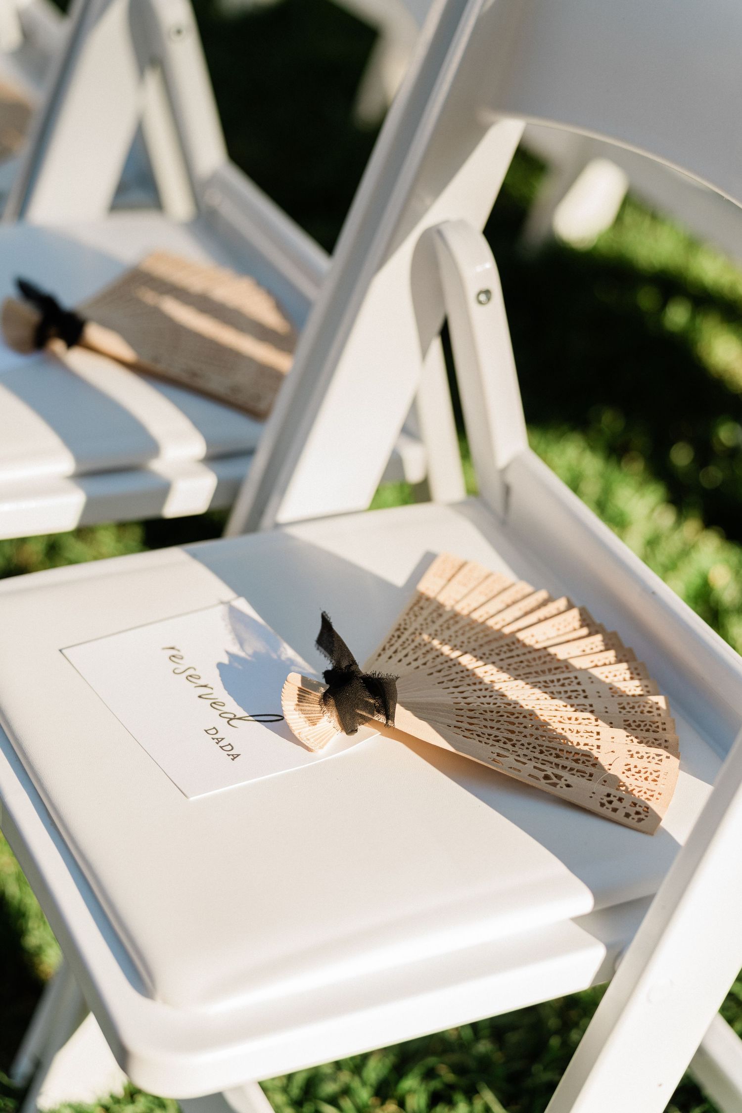 White folding chairs with elegant paper fans and wedding programs placed on seats for an outdoor ceremony.