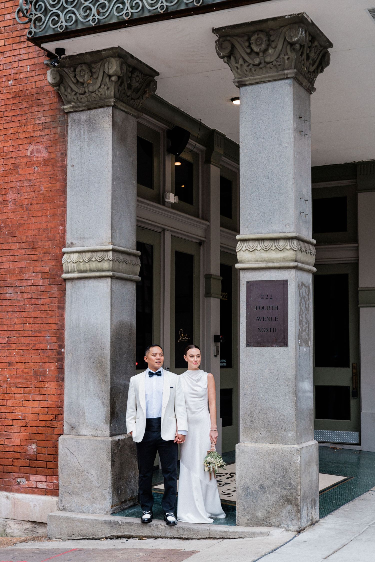 Bride and groom stand between ornate stone columns at a historic brick building entrance on their wedding day.