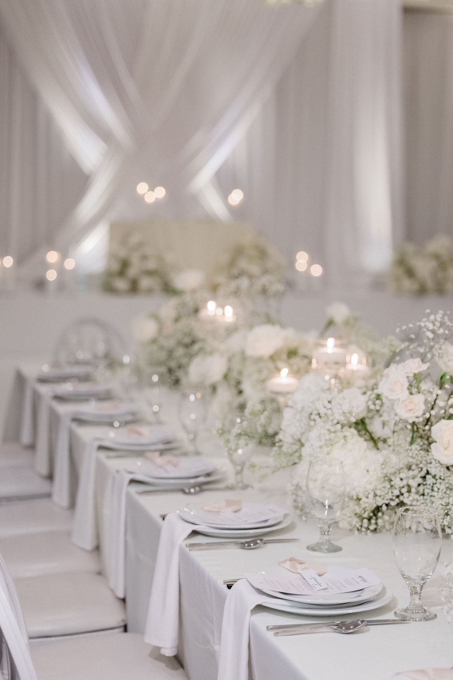 Elegant all-white wedding reception table with floral centerpieces, candles, and draped fabric backdrop.