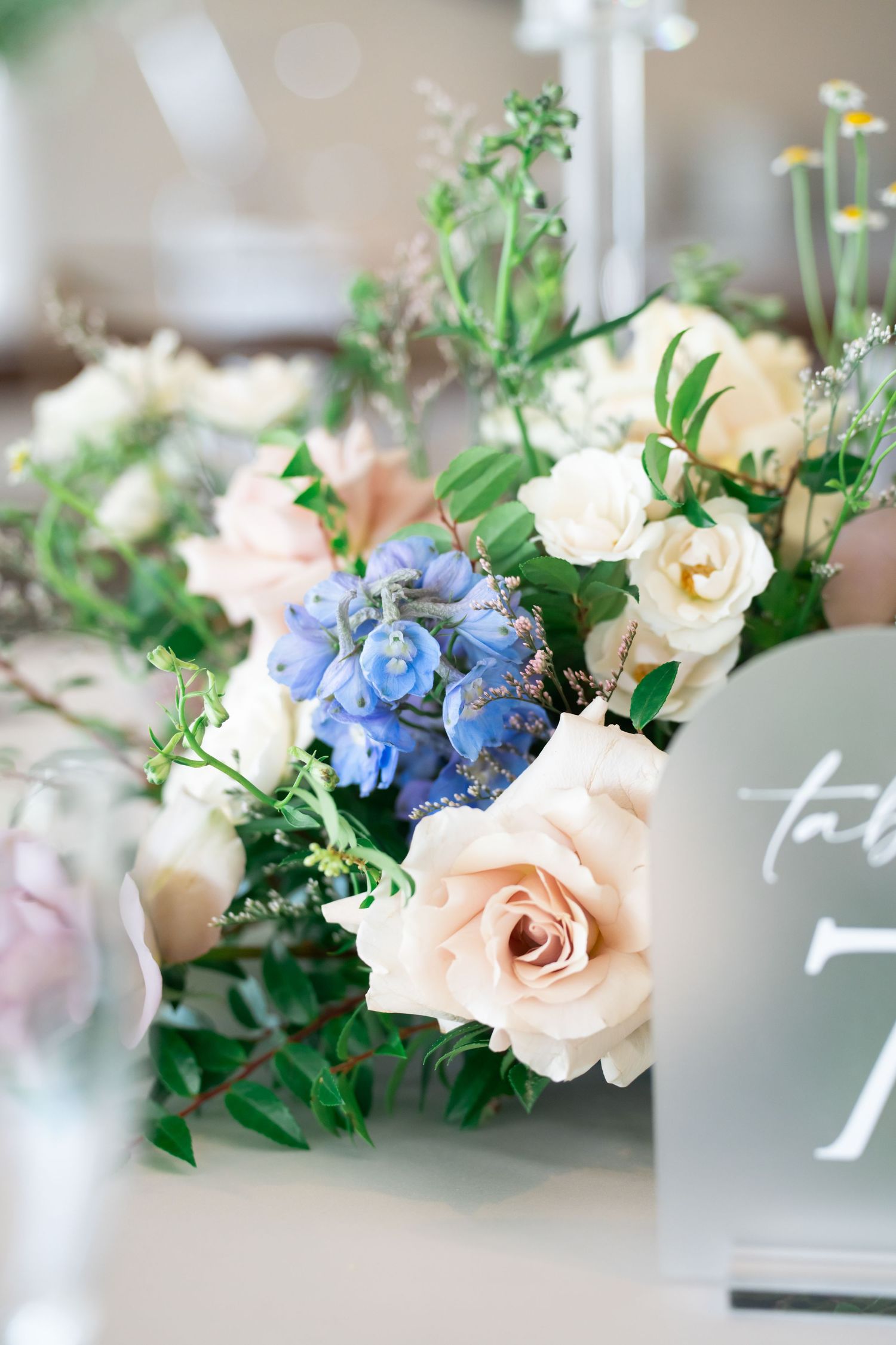 Elegant wedding table centerpiece with blush roses, blue delphiniums, and white blooms beside a table number sign.