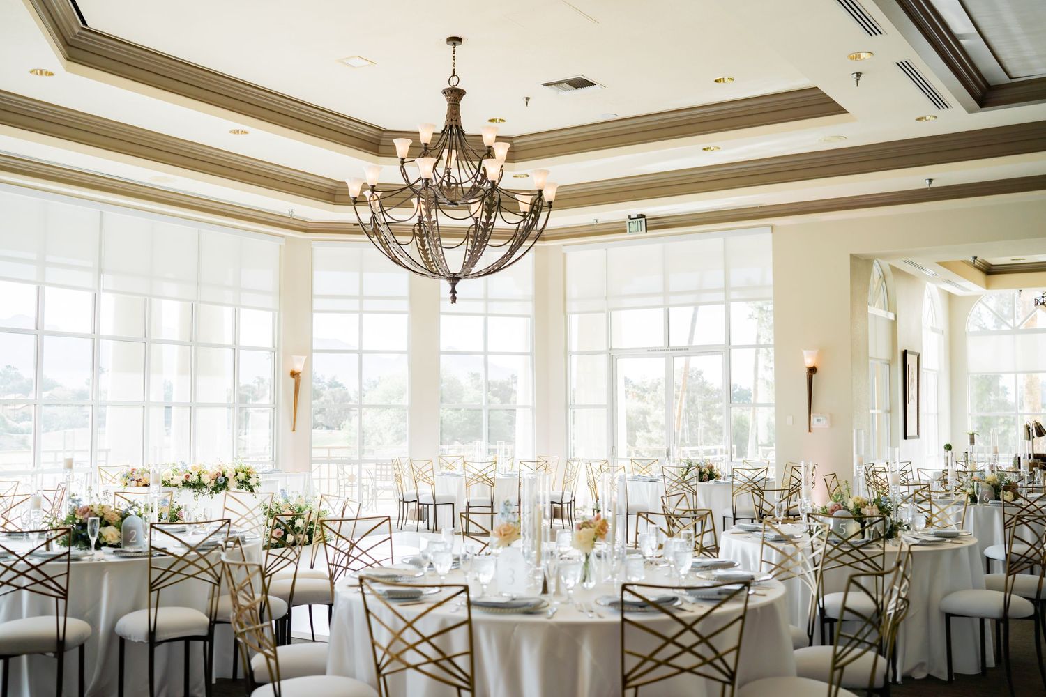 Elegant wedding reception hall with chandelier, gold chiavari chairs, white tablecloths, floral centerpieces, and large windows.